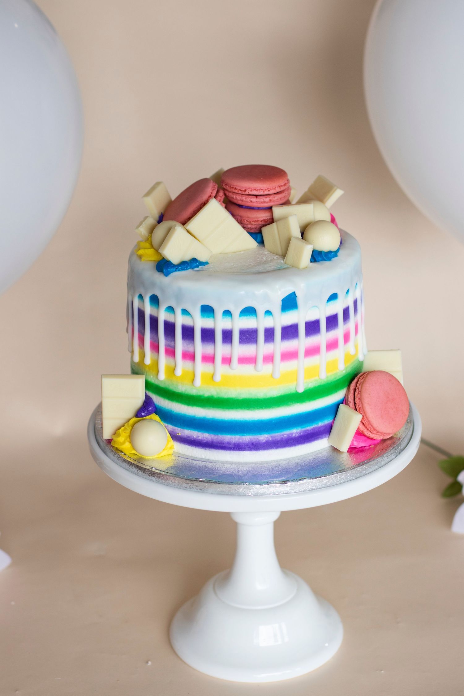 Rainbow striped cake decorated with macarons and colorful details on white cake stand.
