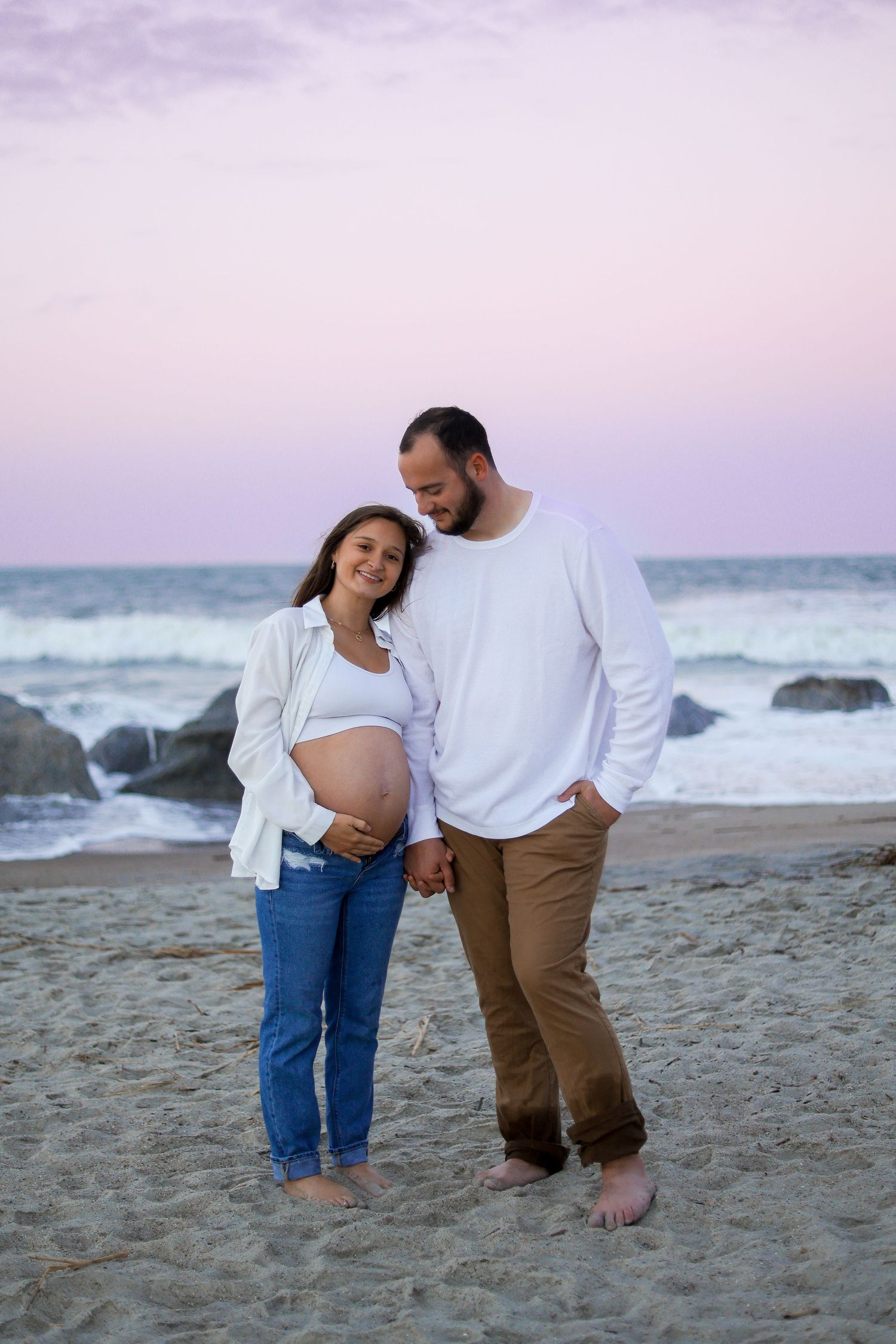 Expectant couple sharing intimate moment on beach with ocean waves and rocks in background.