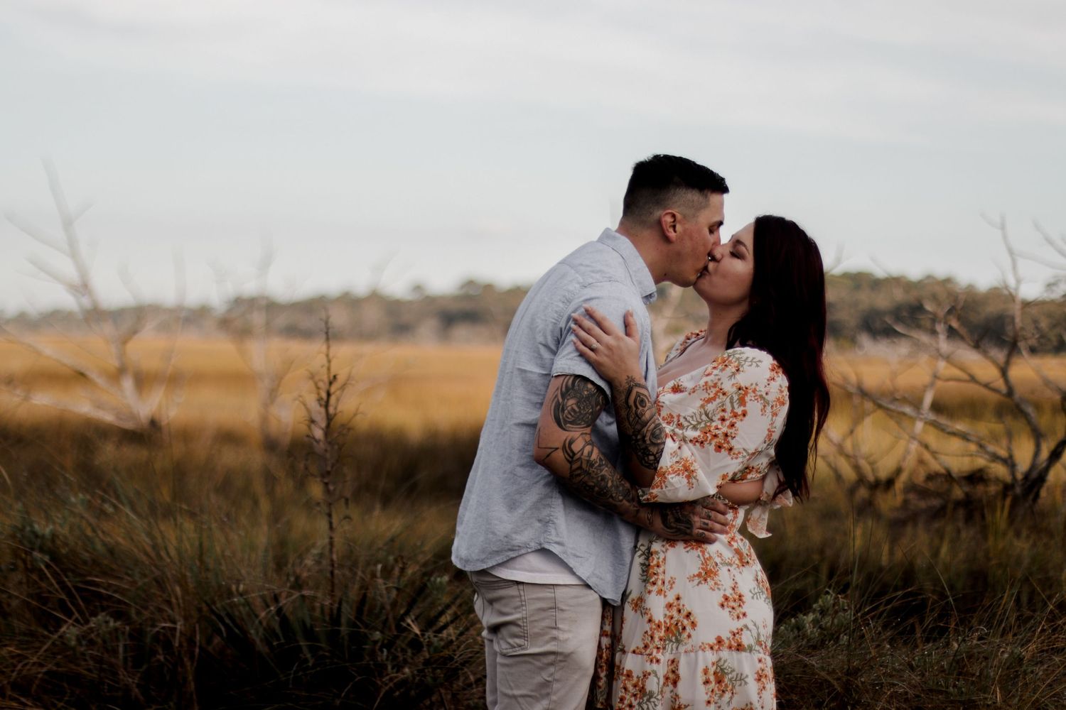 Couple in floral dress and light clothing embracing in golden field at sunset.