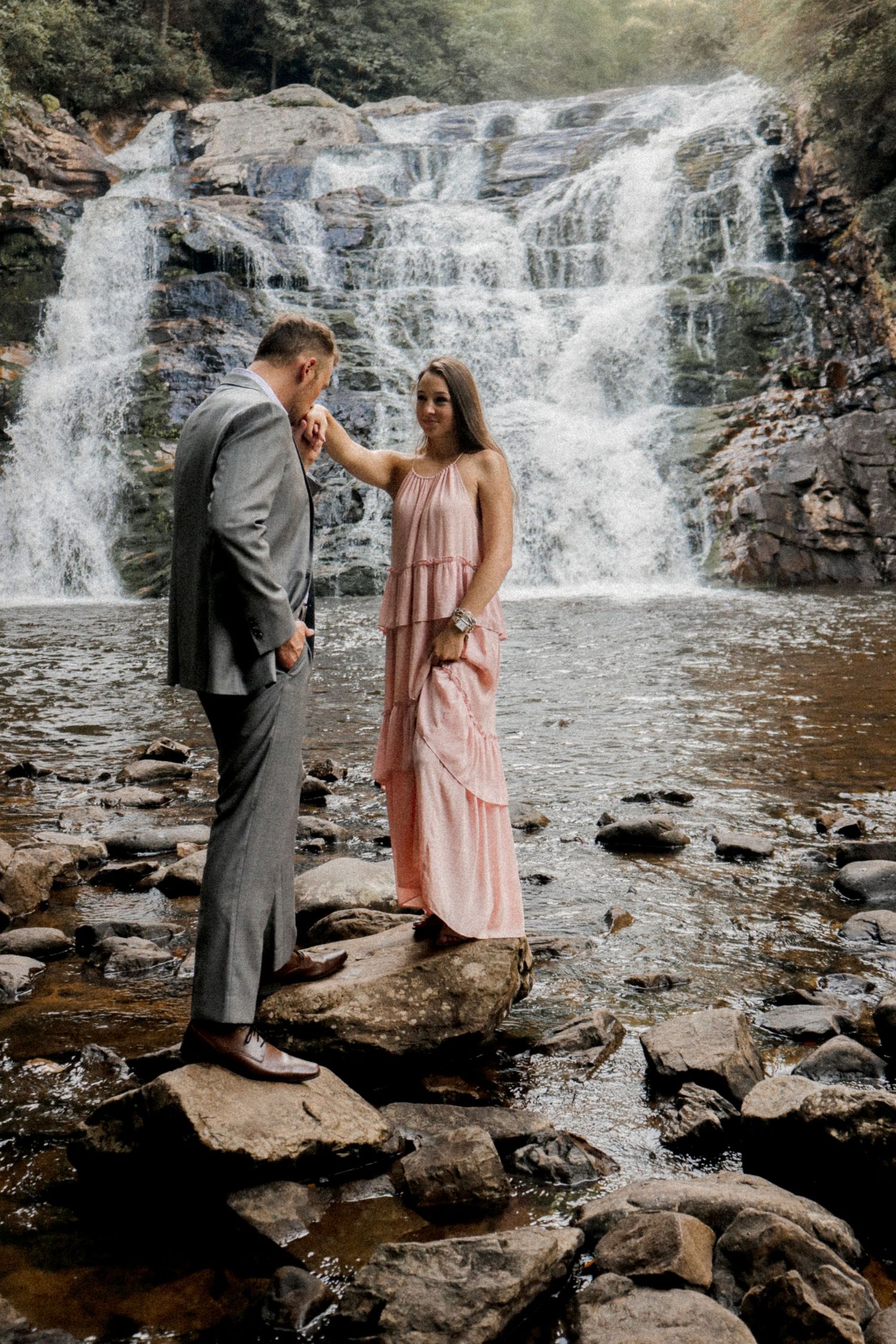 A couple shares a tender moment on rocky terrain with a majestic waterfall backdrop.