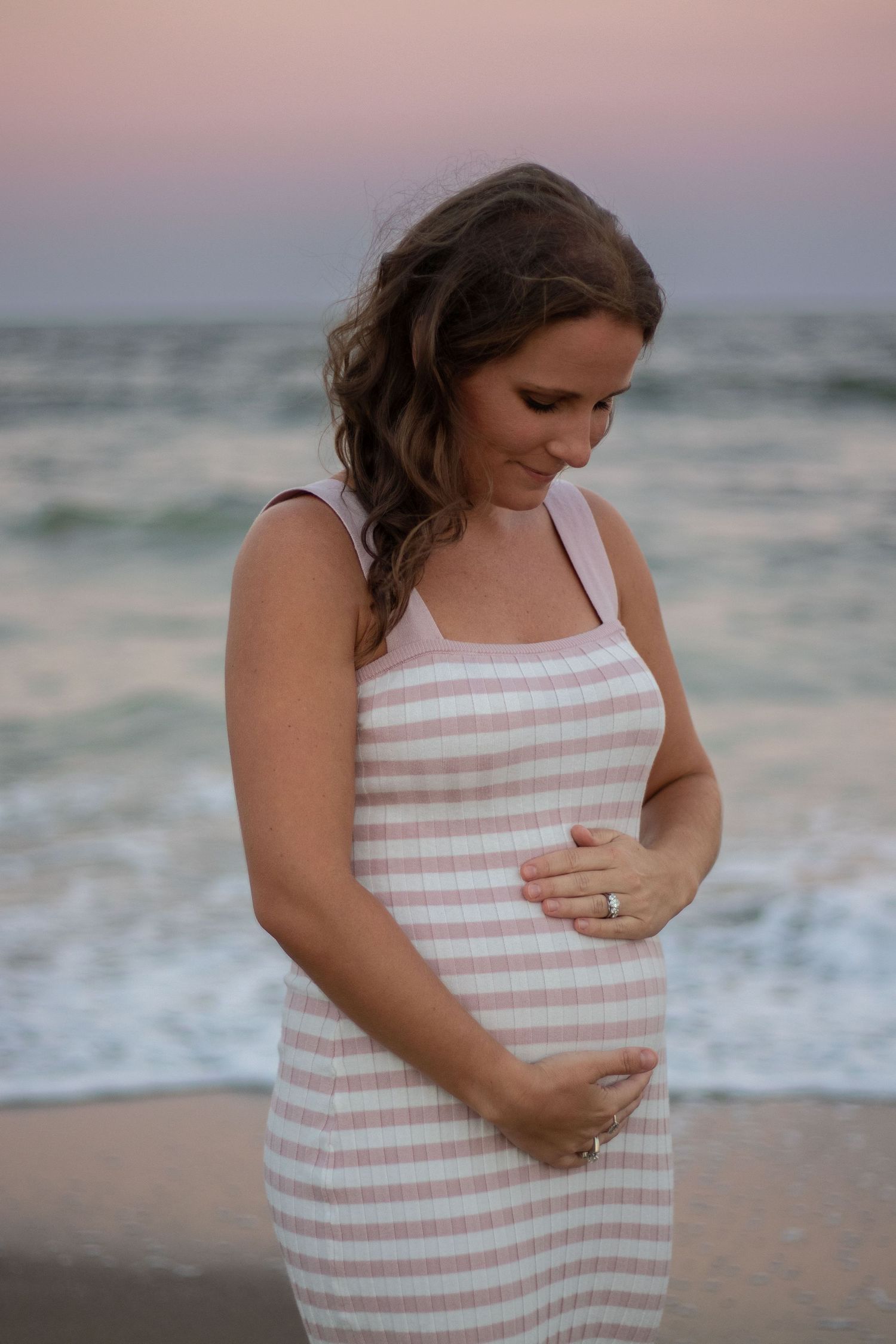 A woman in a striped dress touches her baby bump on the beach at sunset.