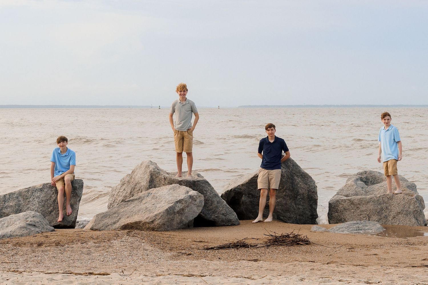 Four people stand on large rocks along a sandy beach at sunset.