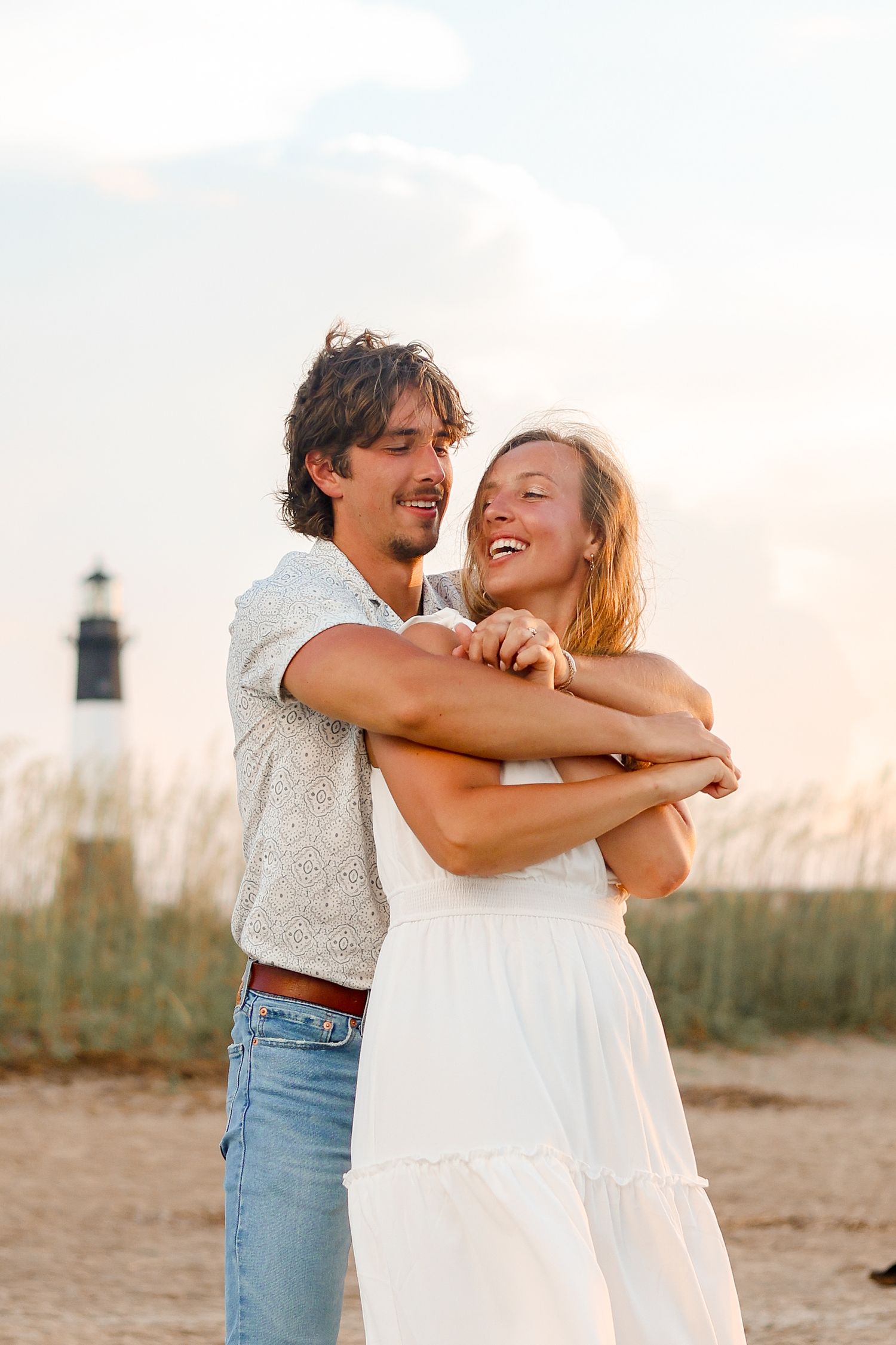 Happy couple embracing by the lighthouse during a beach photoshoot at sunset.