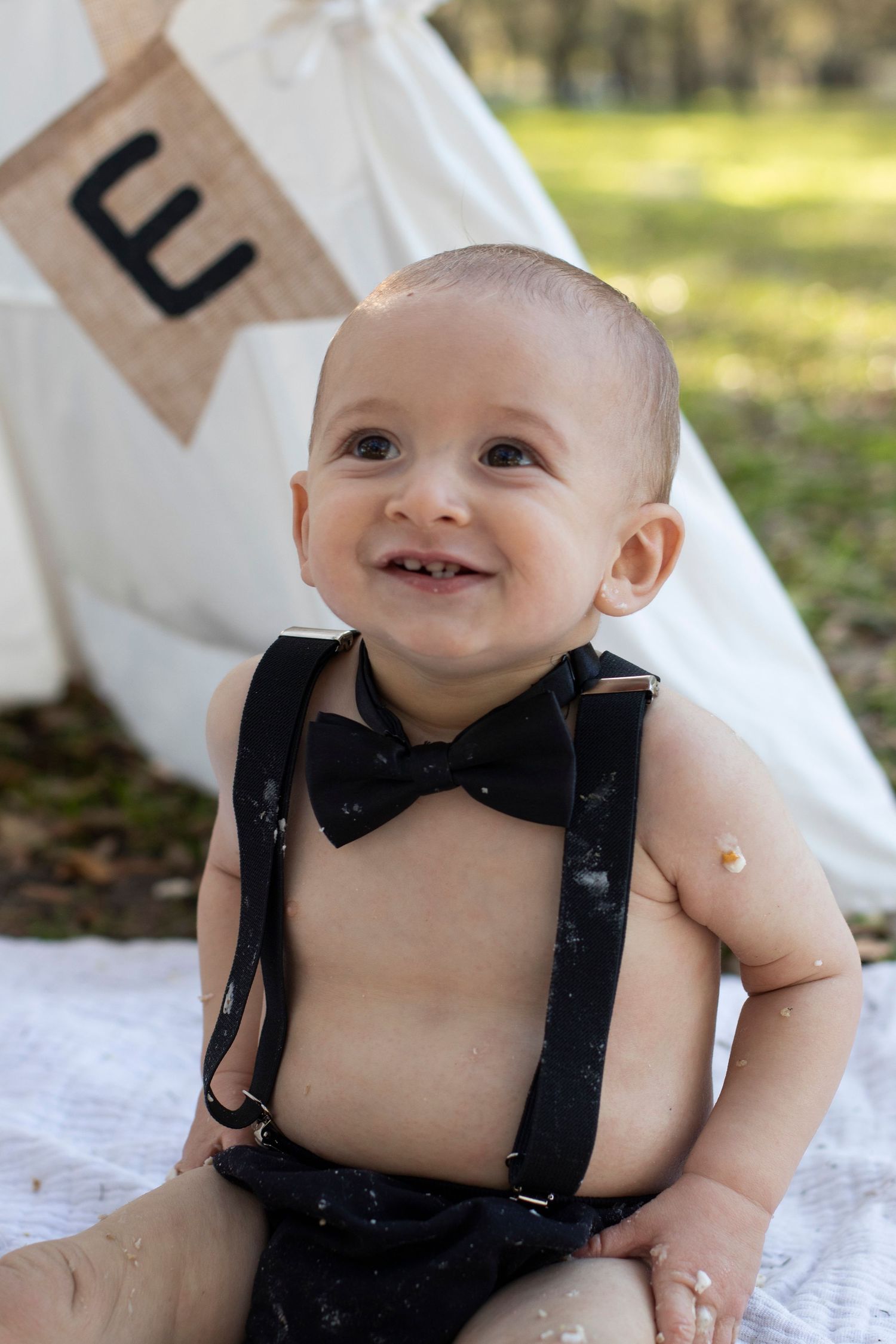 Baby in black suspenders smiling during outdoor photoshoot.
