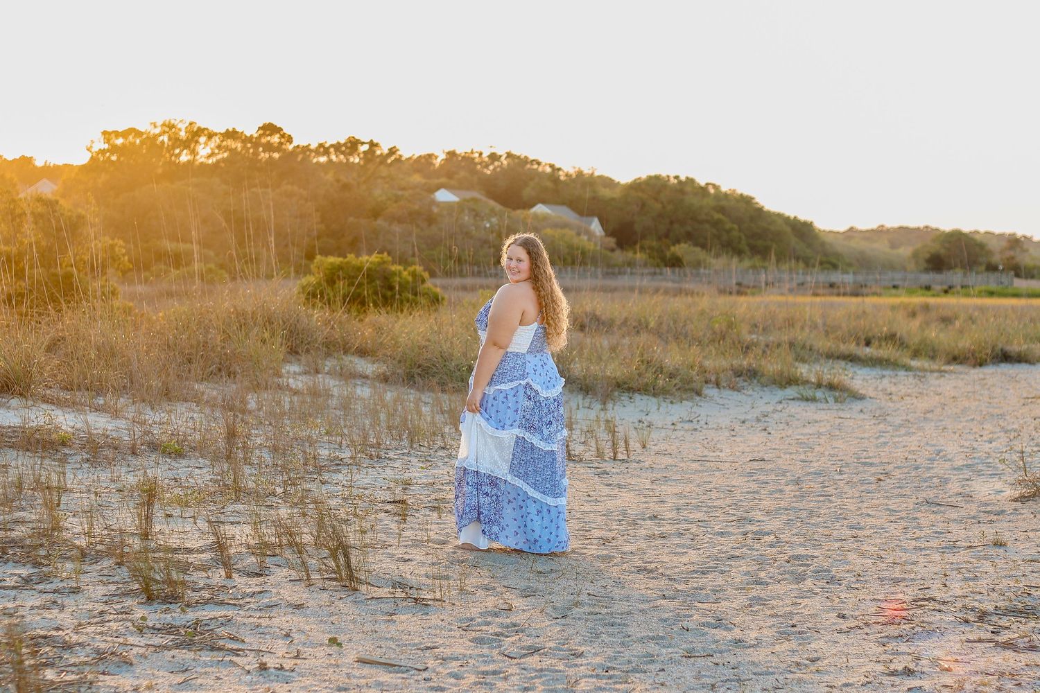 Woman in a flowing blue dress stands on a sandy beach during golden hour sunset.