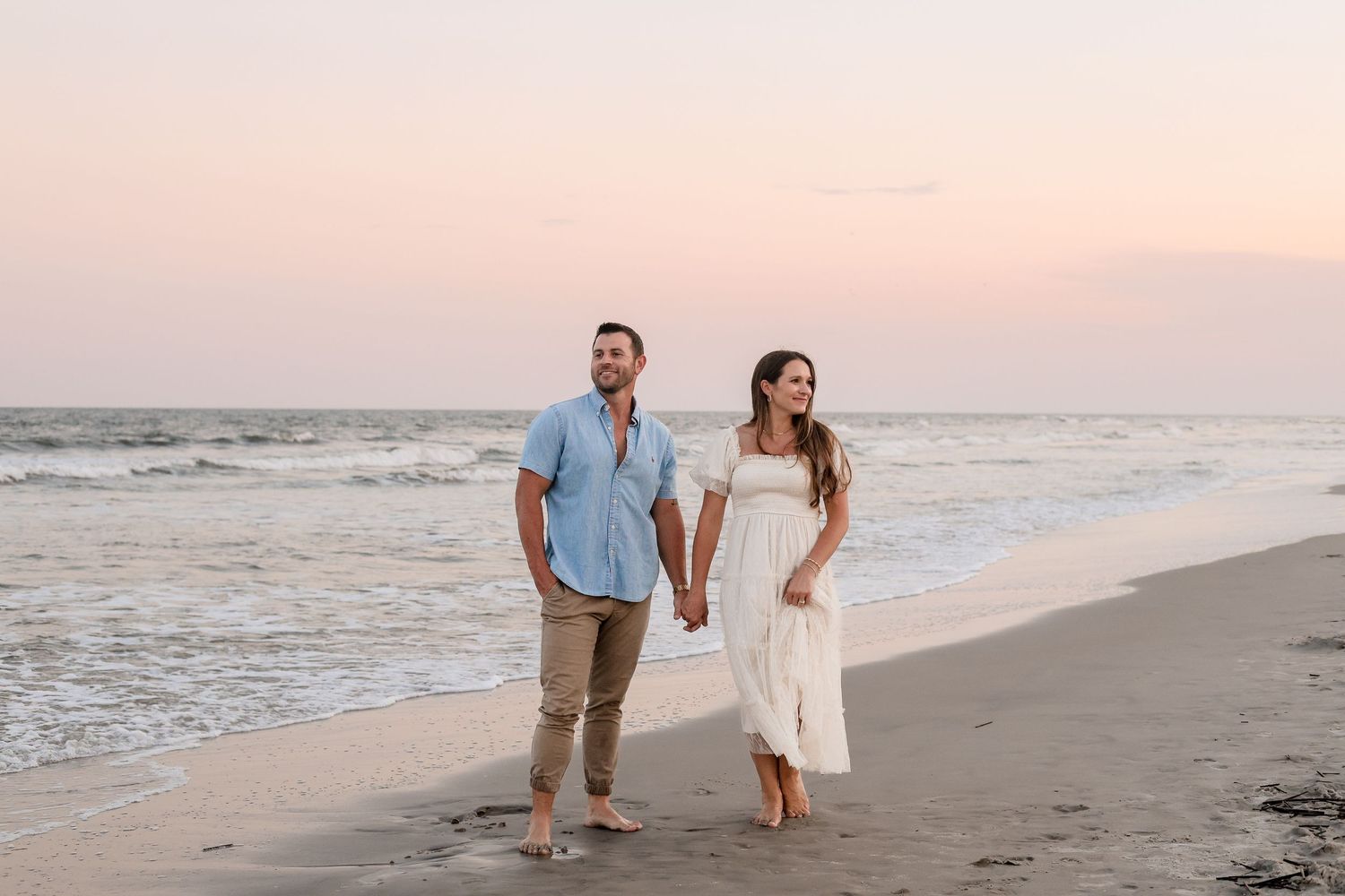 A couple walks hand in hand along a serene beach at sunset with waves rolling onto the shore.