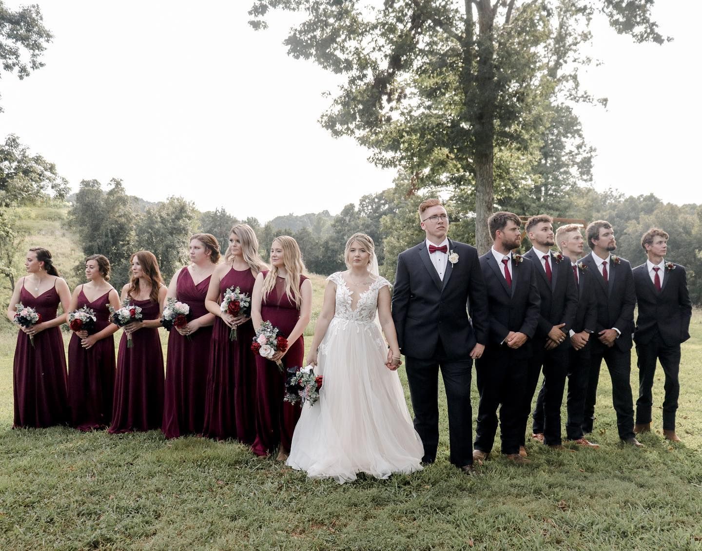A wedding party in burgundy dresses and black suits poses together outdoors.