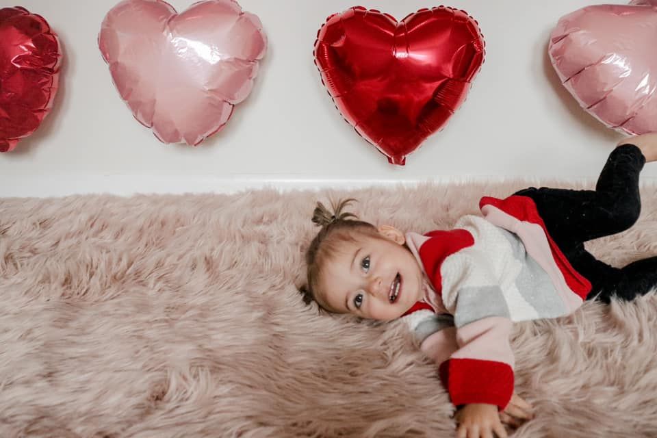 Toddler laying on plush carpet surrounded by pink and red heart-shaped balloons.