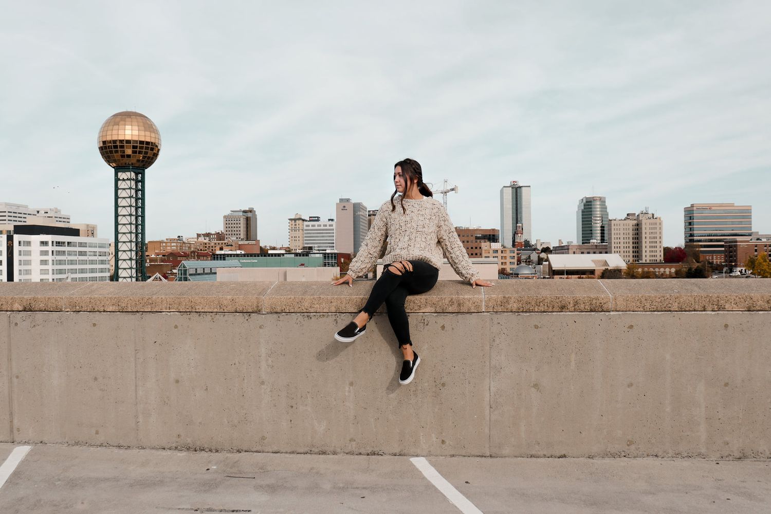 Woman sitting on concrete wall overlooking city skyline with distinctive spherical tower.