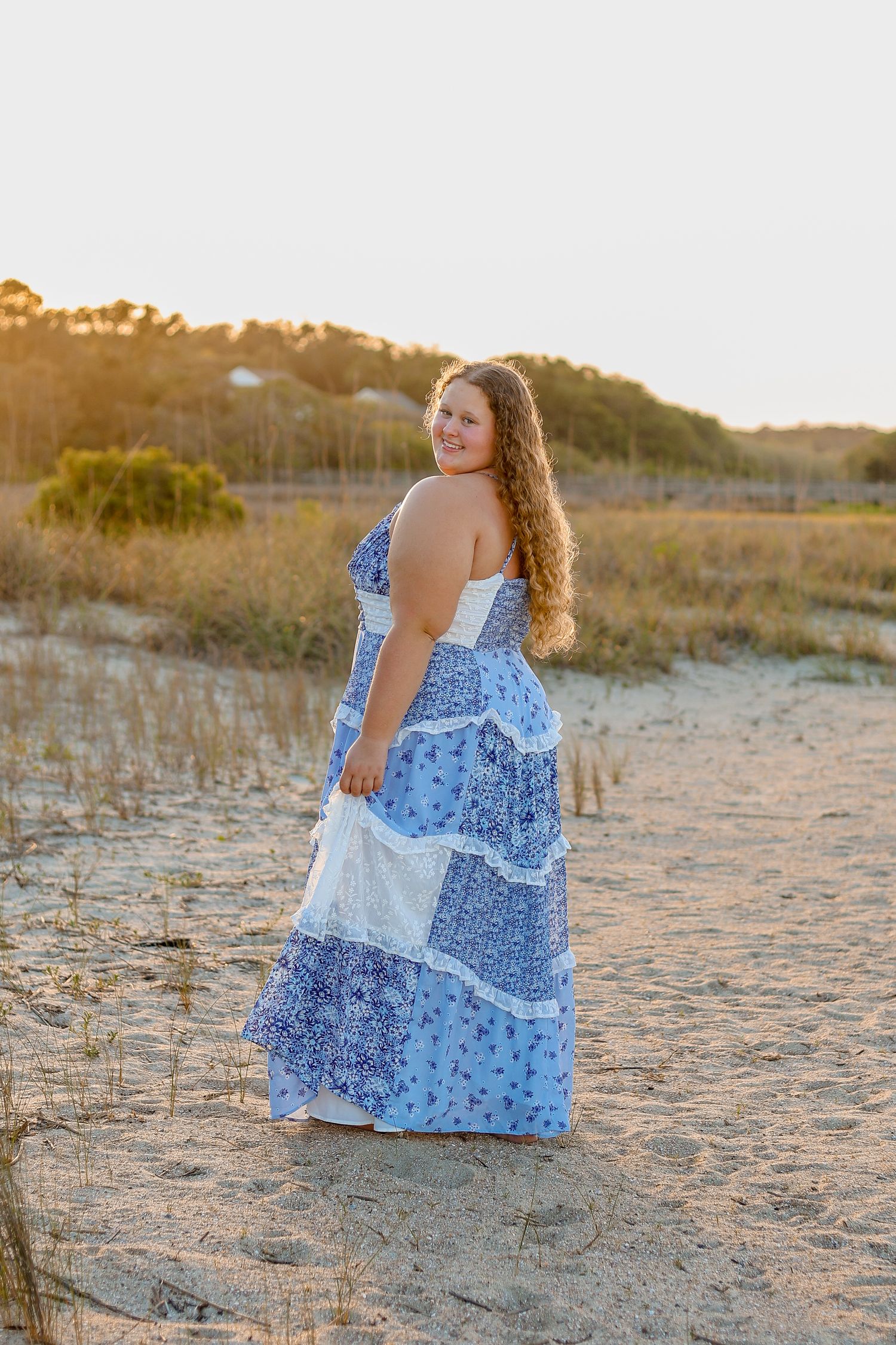 Woman in blue patchwork dress sits on sandy beach during golden sunset hour.
