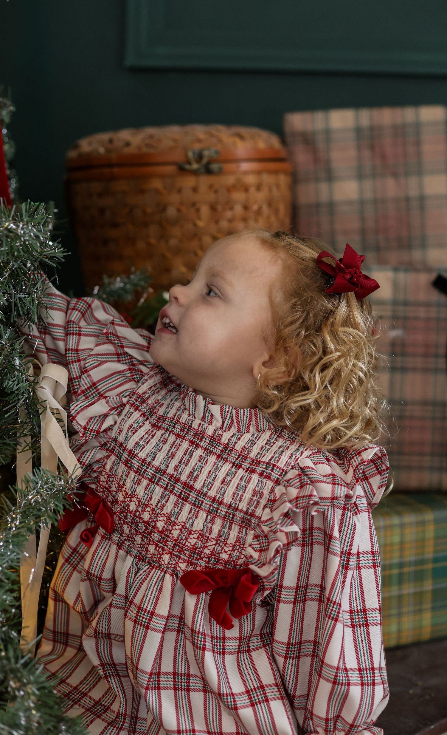 A child wearing a red and white plaid dress with ruffles and a red bow smiles while looking up during holiday decorating.