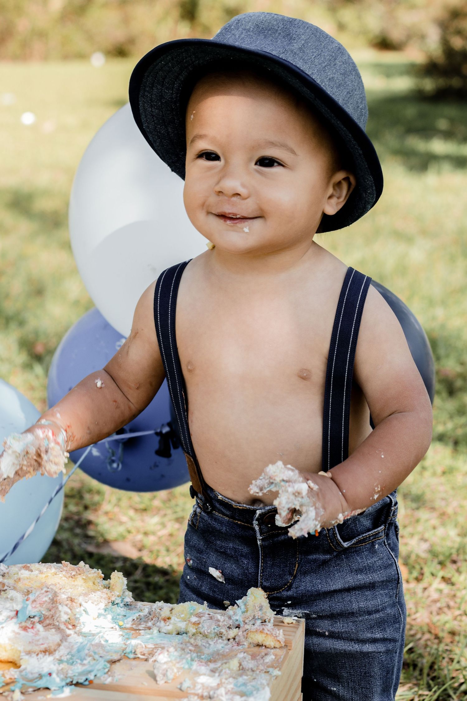 Toddler in overalls and hat plays with cake smash setup in sunny outdoor setting.