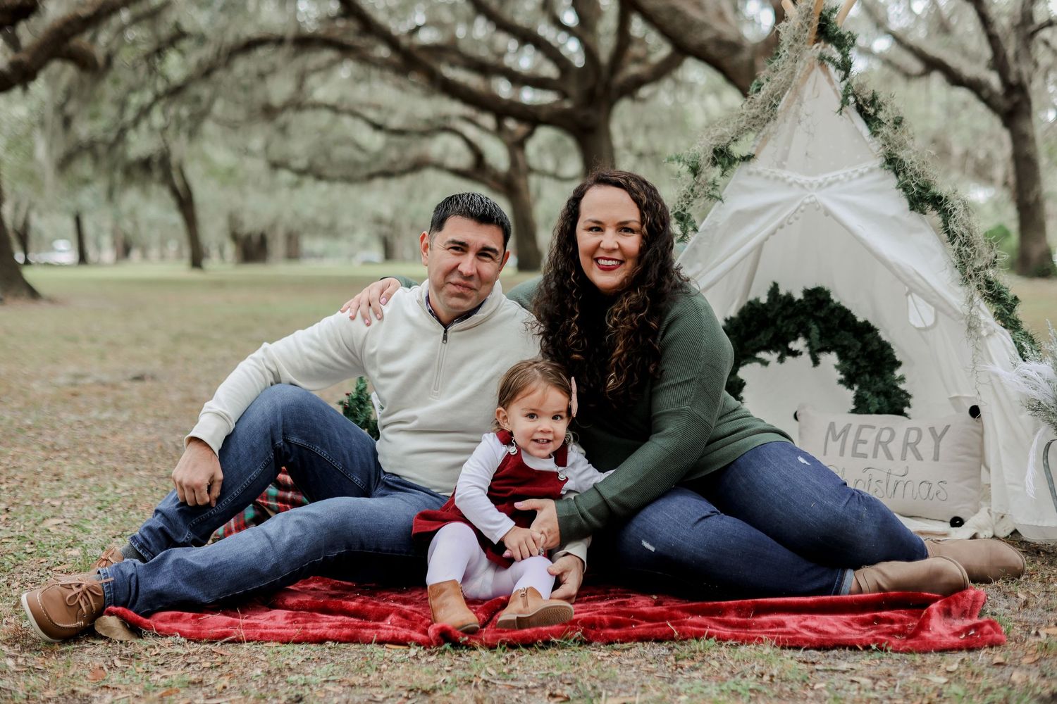 A family sits together on a red blanket next to a white teepee tent outdoors.