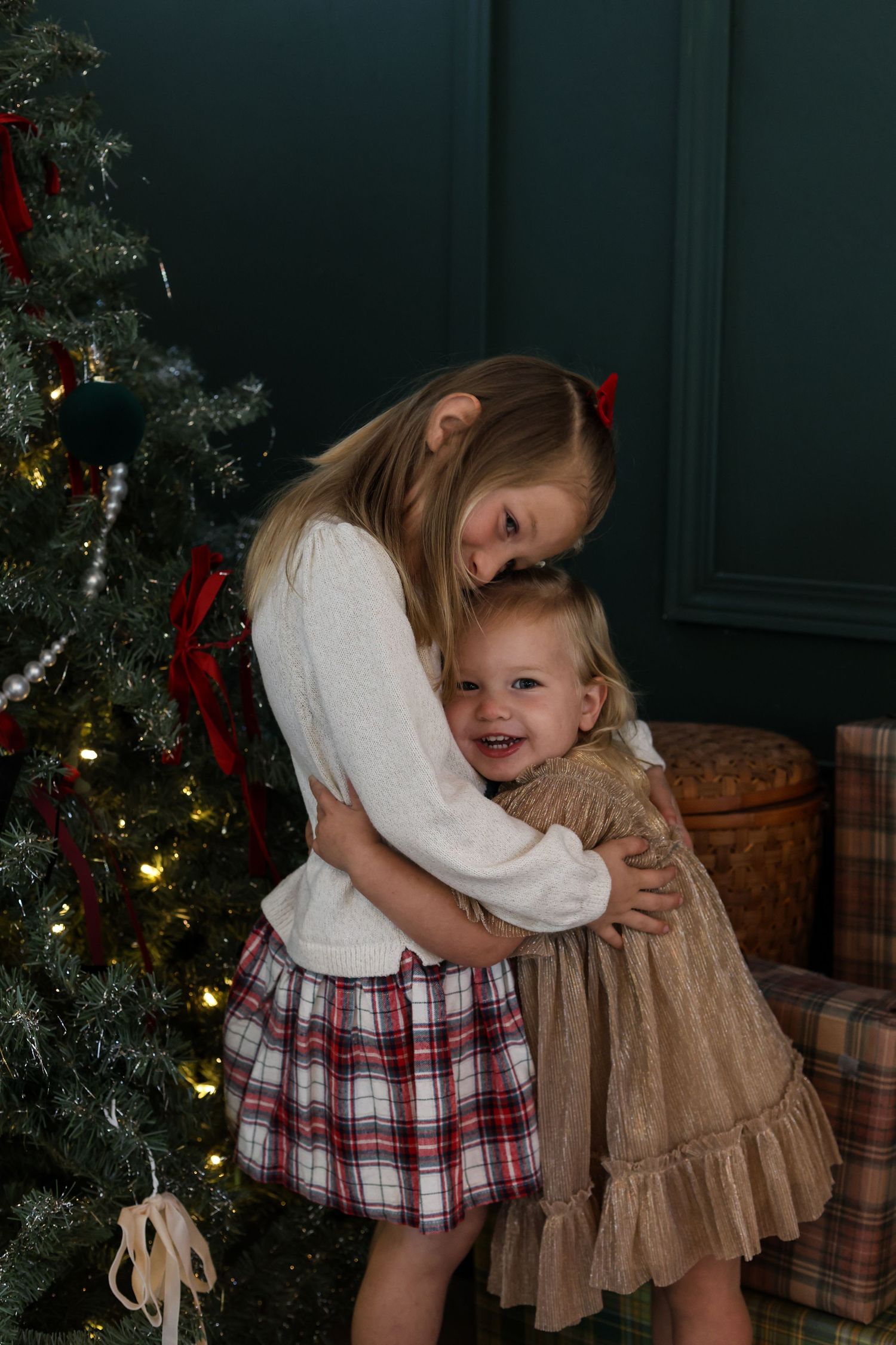 Two children embrace near a decorated Christmas tree with plaid and ruffled dresses in a cozy indoor setting.