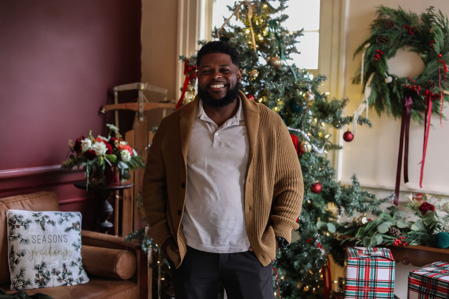 Smiling man in a tan cardigan stands beside a decorated Christmas tree with a festive wreath, gifts, and a holiday pillow in a cozy room.