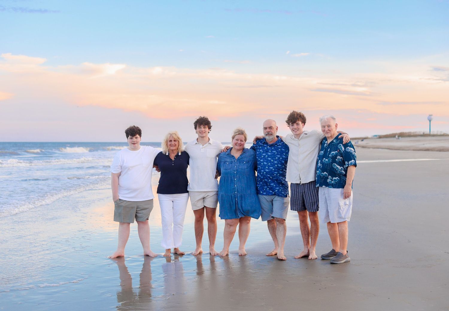 Group wearing coordinated blue and white outfits poses together on sandy beach at sunset.