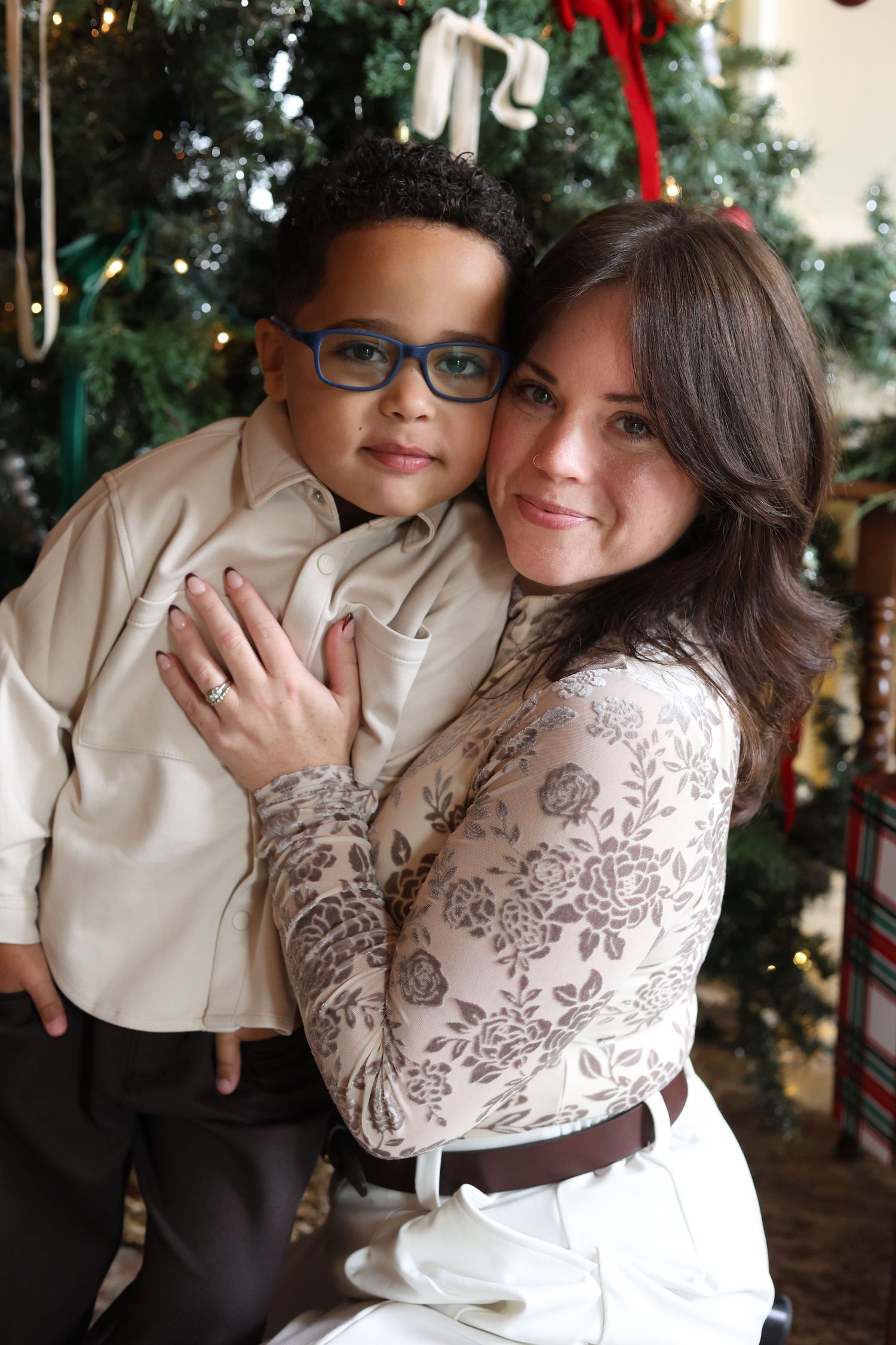 A smiling woman in a floral lace top hugs a young boy in glasses and a cream shirt beside a decorated Christmas tree.