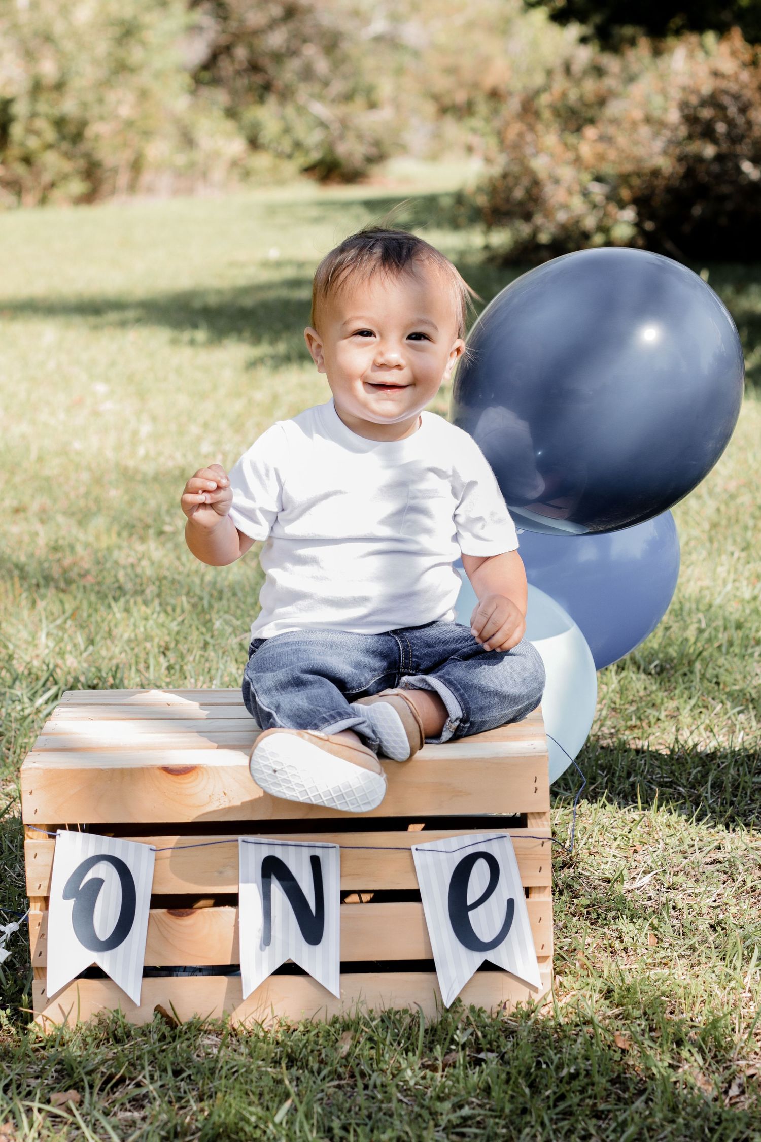 Baby in white outfit sitting on wooden crate with blue balloons and ONE banner outdoors.