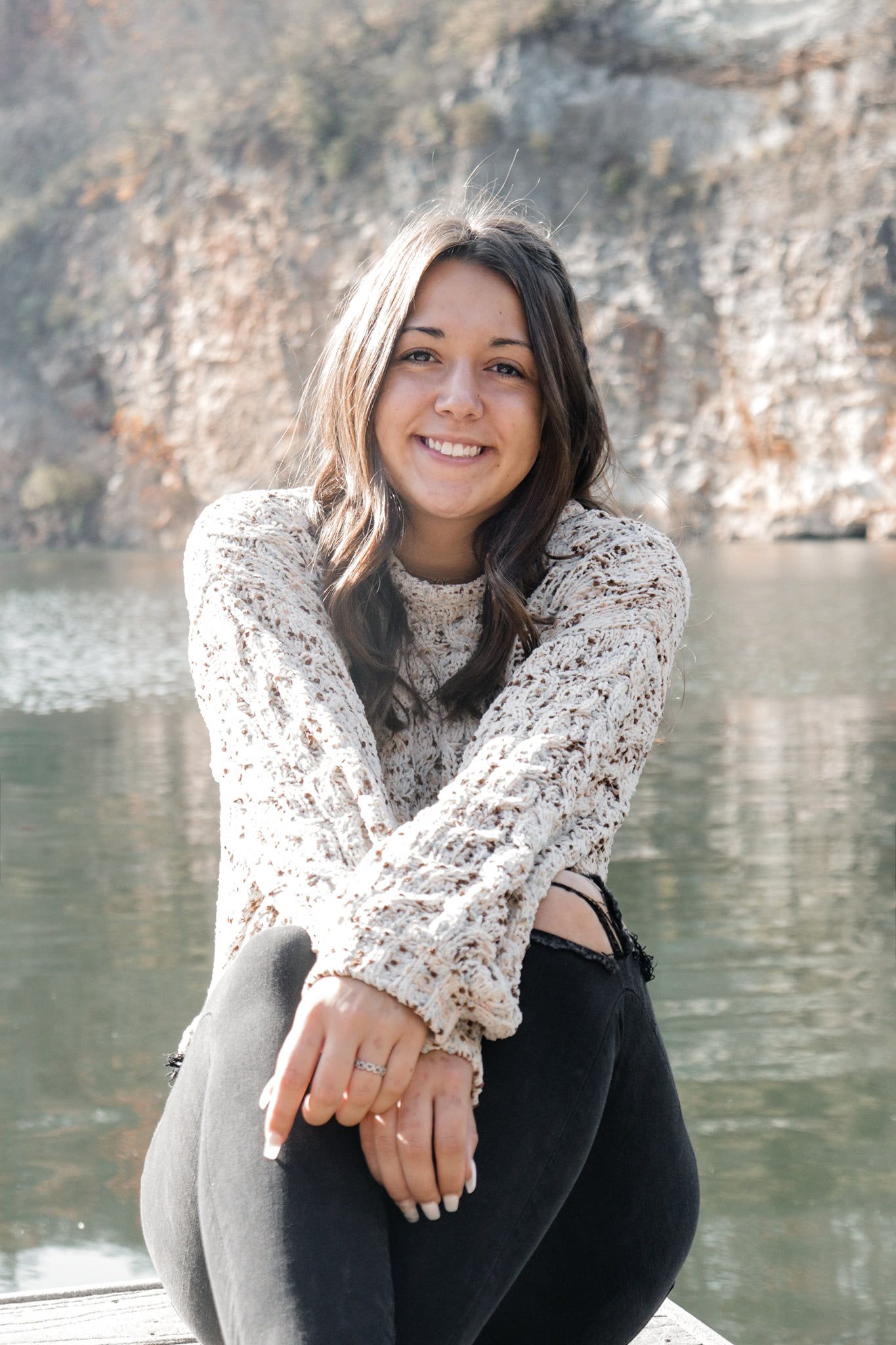 Portrait by a serene lake with rocky cliffs wearing a cozy white knit sweater.
