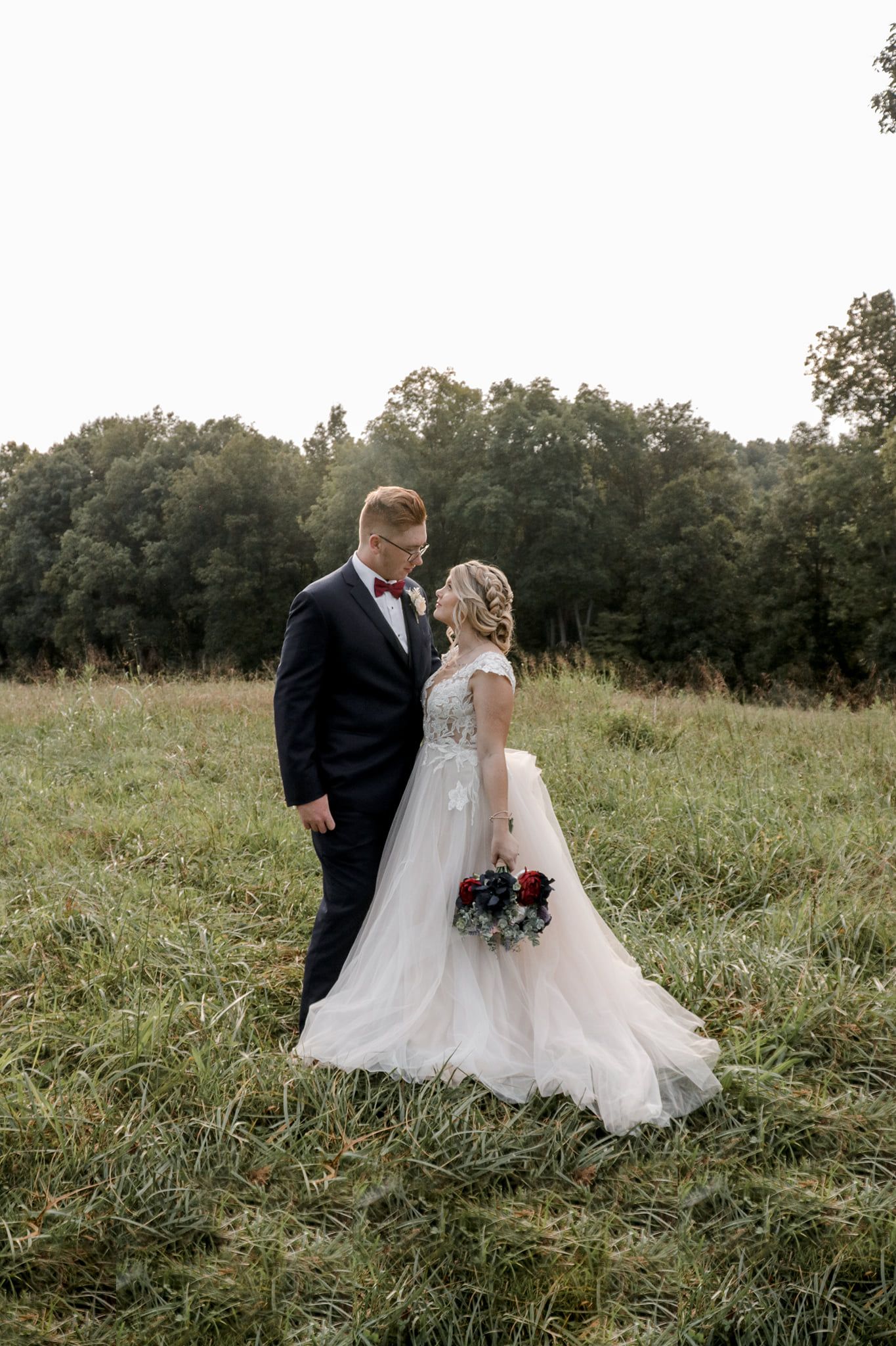 Wedding couple standing in field with bride holding purple bouquet at sunset.