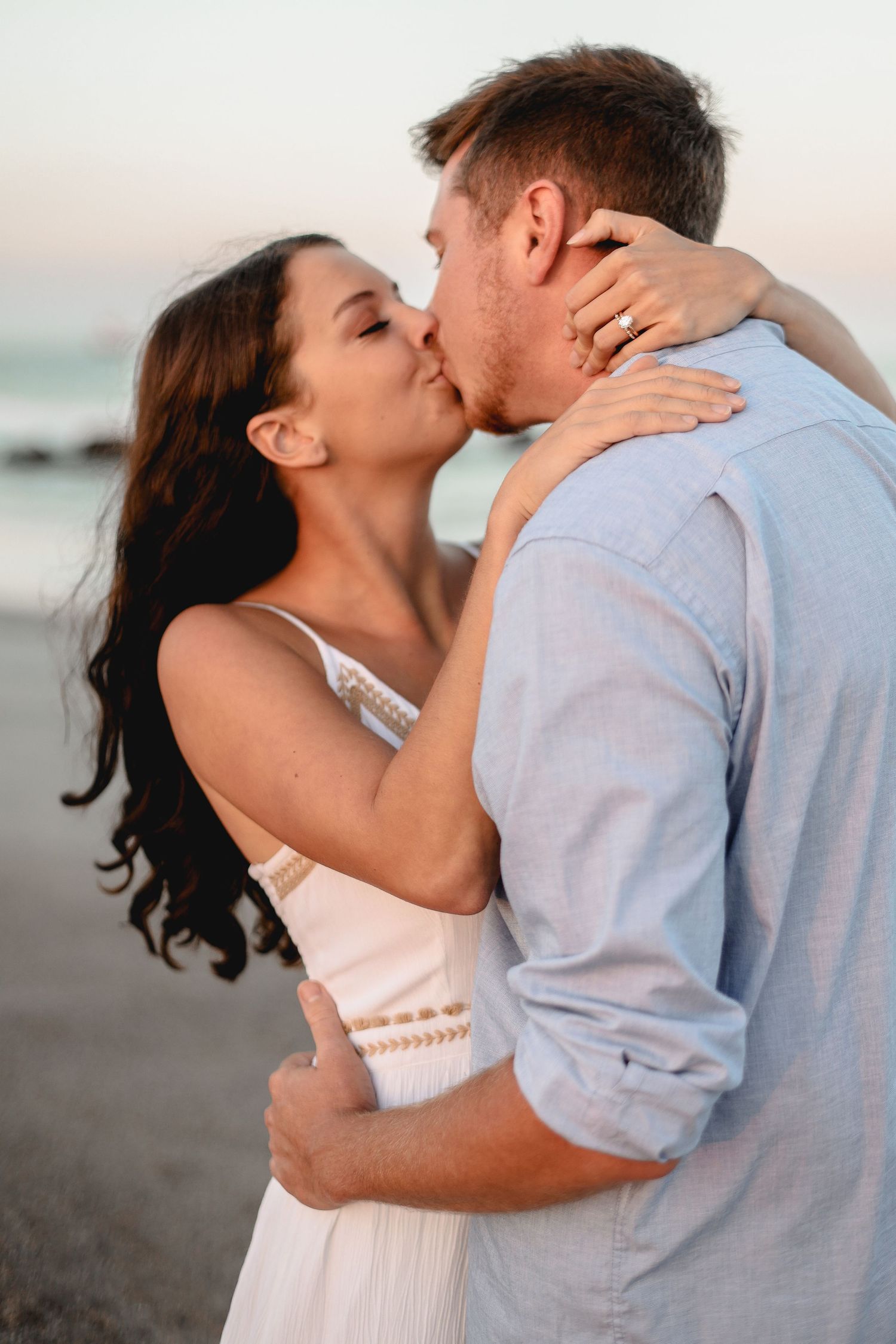 A romantic kiss shared between a newly engaged couple on the beach at sunset.