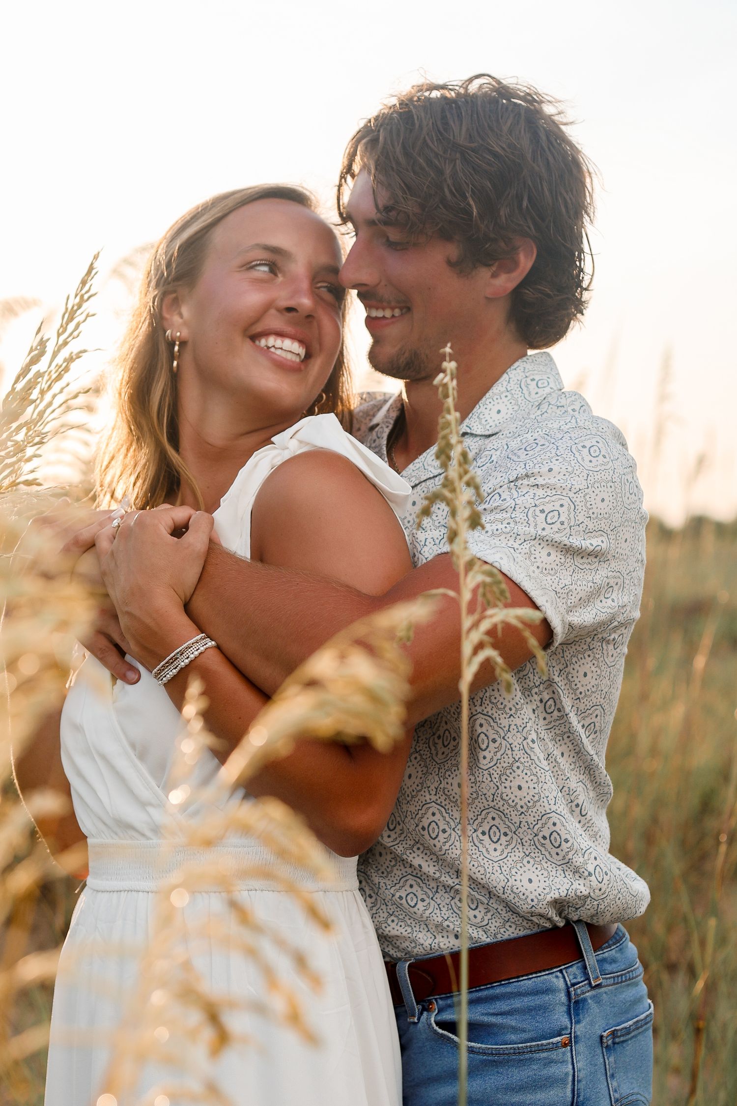 Romantic couple embracing in a field of tall grass during golden hour at sunset.