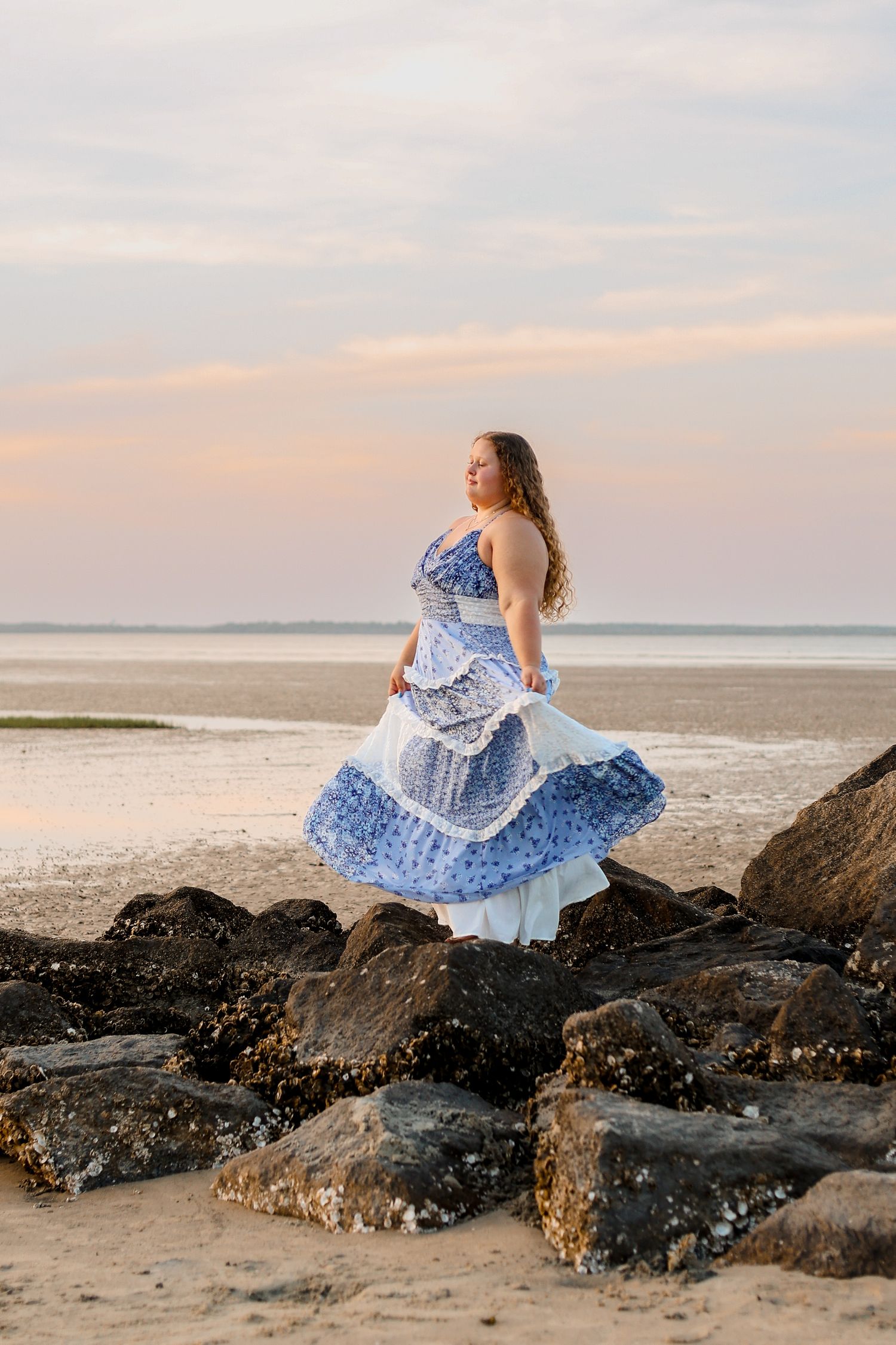 Person in a flowing blue and white tiered dress poses gracefully on coastal rocks during sunset at the beach.