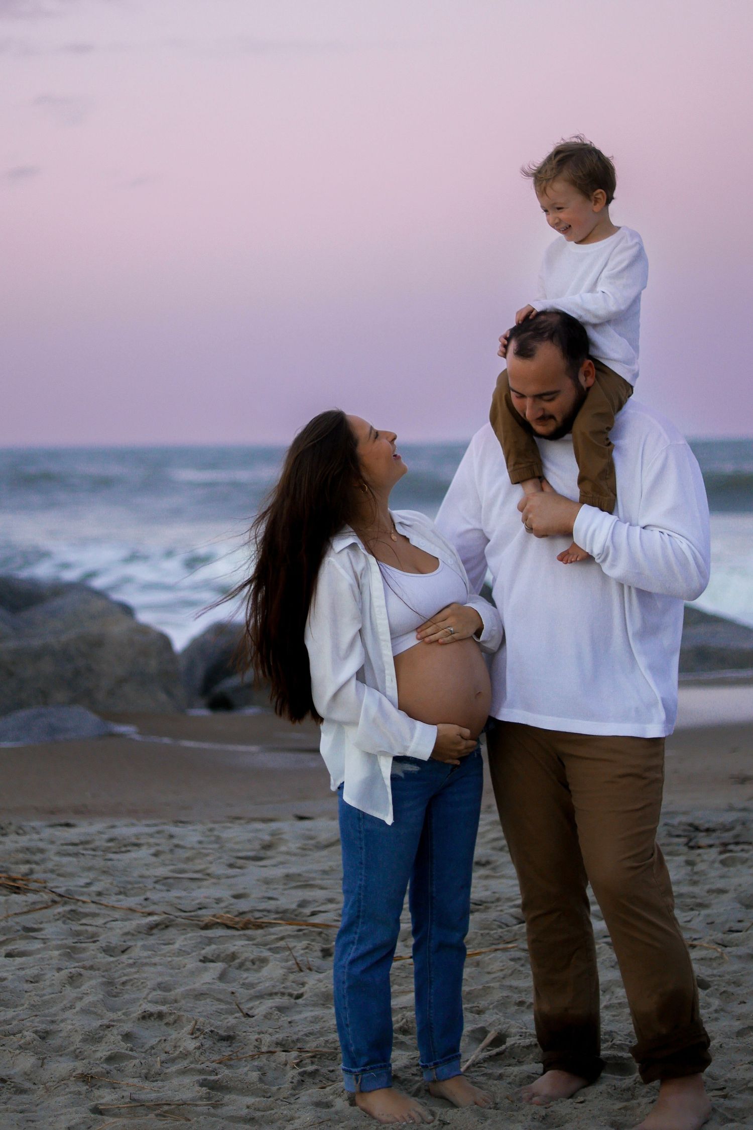 Family enjoying sunset beach moment with toddler on shoulders and waves crashing behind them.