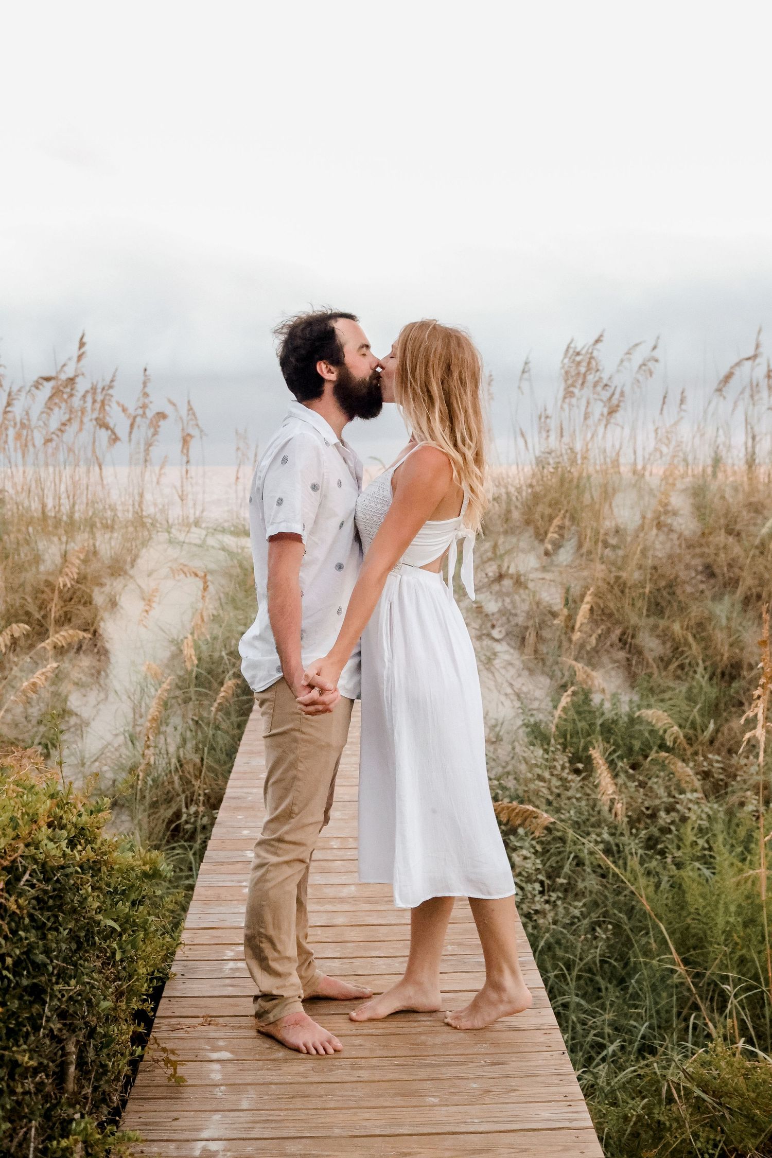 Romantic couple sharing a tender moment on a wooden boardwalk surrounded by tall grass at sunset.