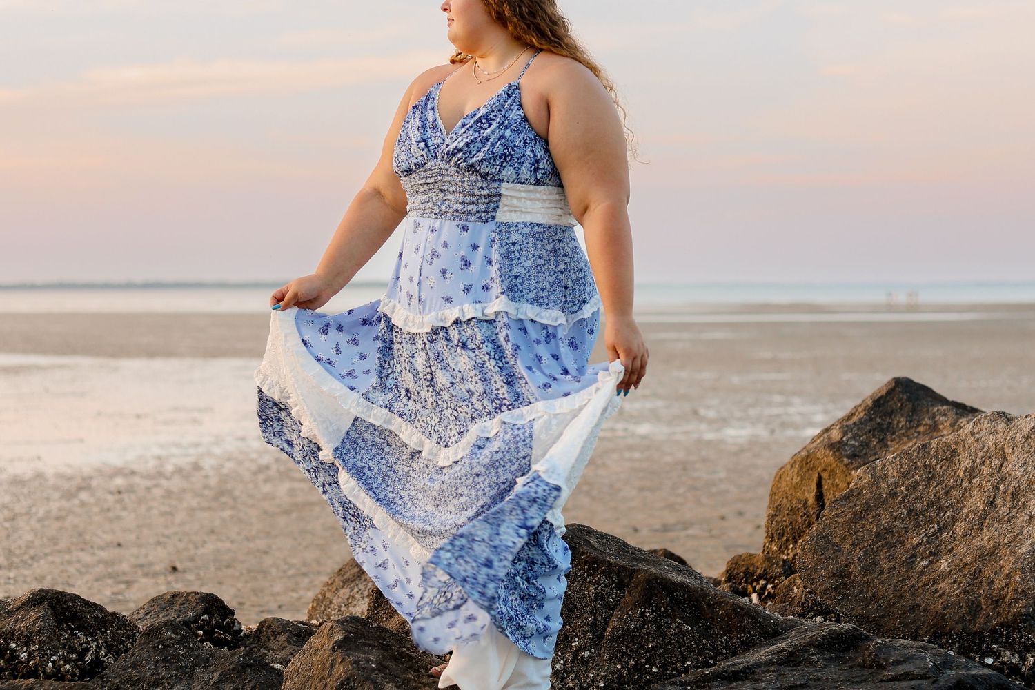 A woman in a flowing blue and white patterned maxi dress stands gracefully on coastal rocks at sunset.