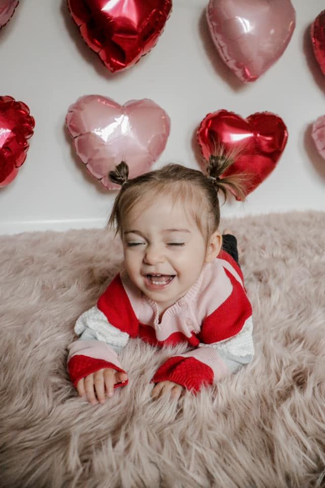 Toddler laying on plush carpet with red and pink heart balloons decorating the wall behind.