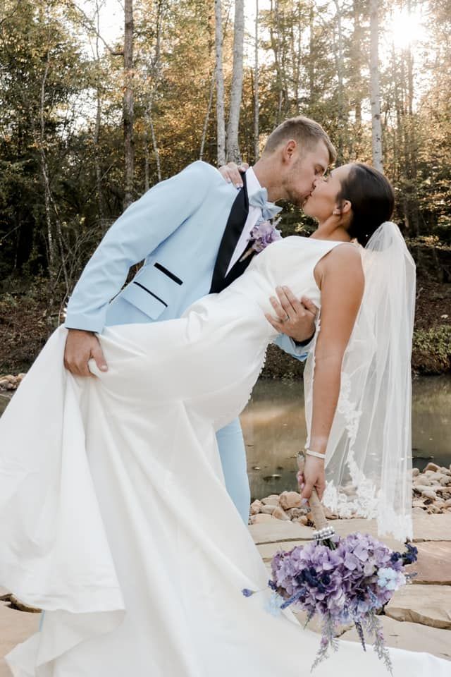 Romantic wedding couple sharing a dip kiss outdoors with purple flowers in a wooded setting during sunset.