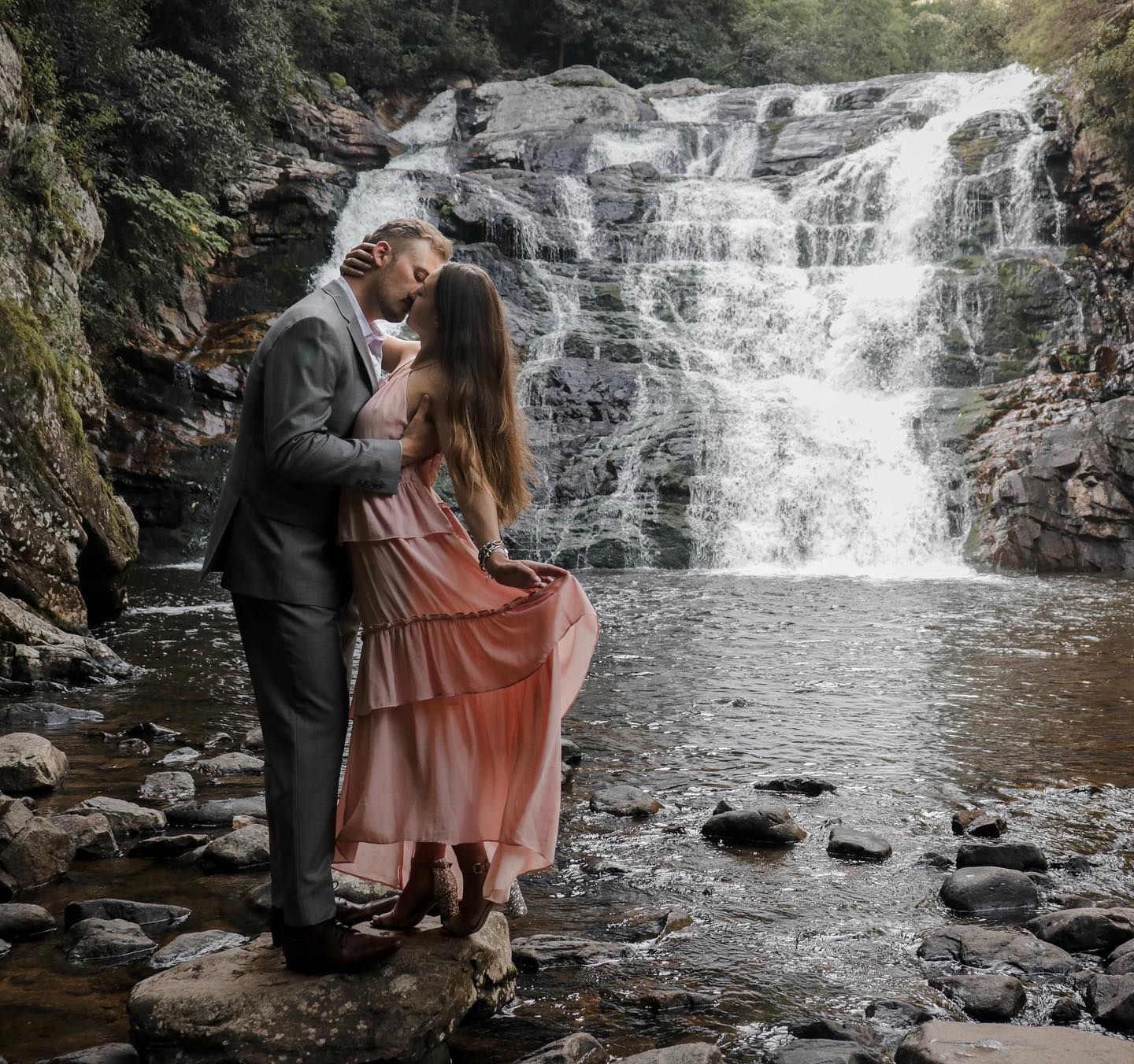 A couple poses romantically by a rushing waterfall in nature.