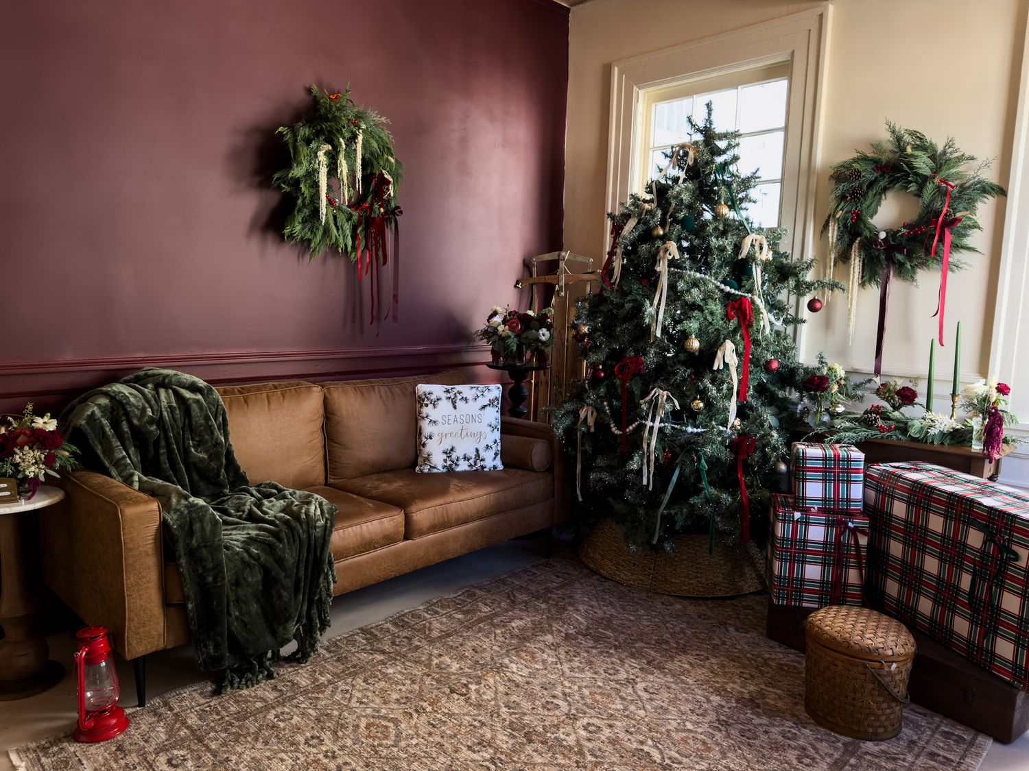Cozy living room decorated for Christmas with a lit tree, evergreen wreaths, plaid gift boxes, a tan leather sofa and green knit throw.