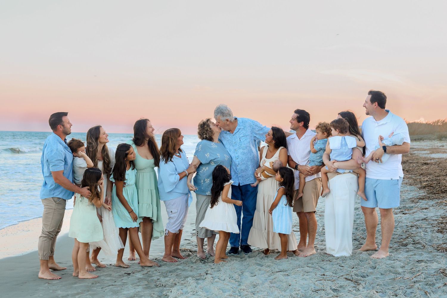Extended family group wearing coordinated blue and white outfits stands together on beach at sunset.