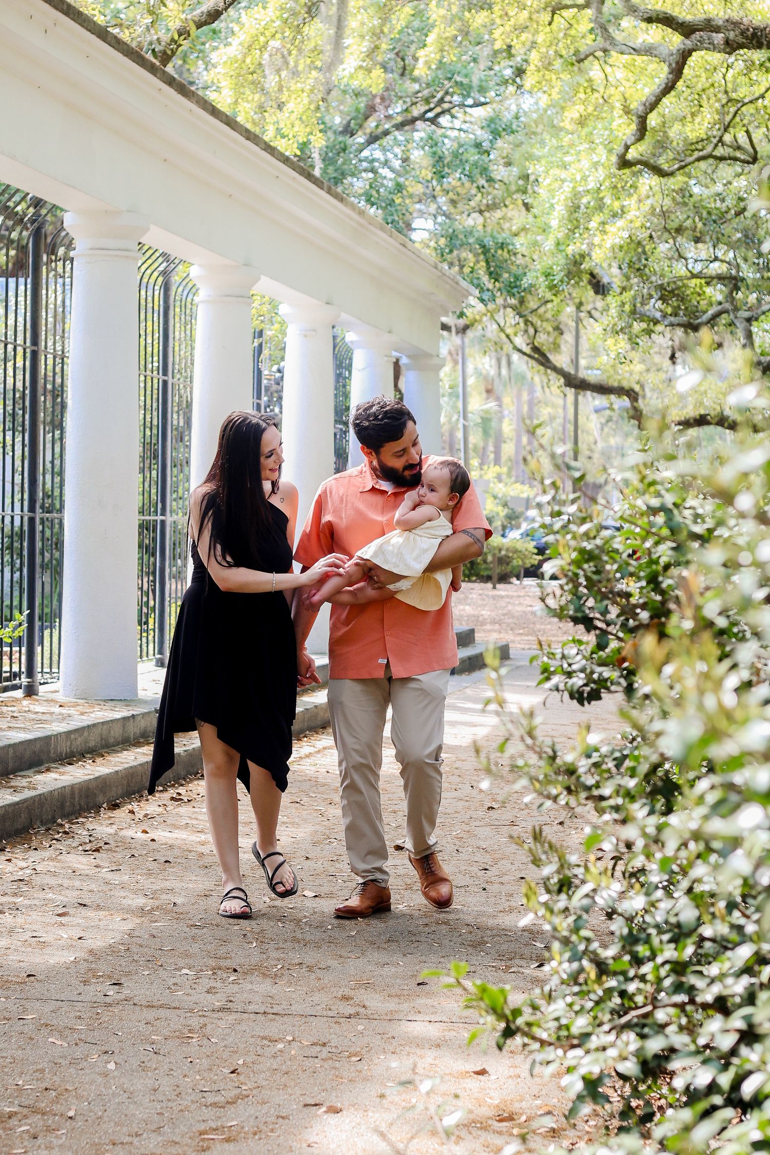 A couple walking with their newborn baby along a columned walkway in a garden setting.