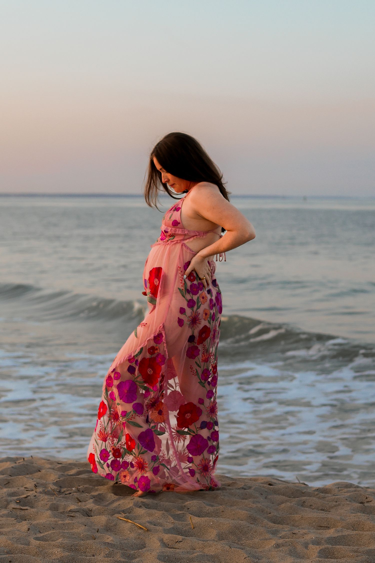 Pregnant woman in floral maxi dress poses on beach at sunset.