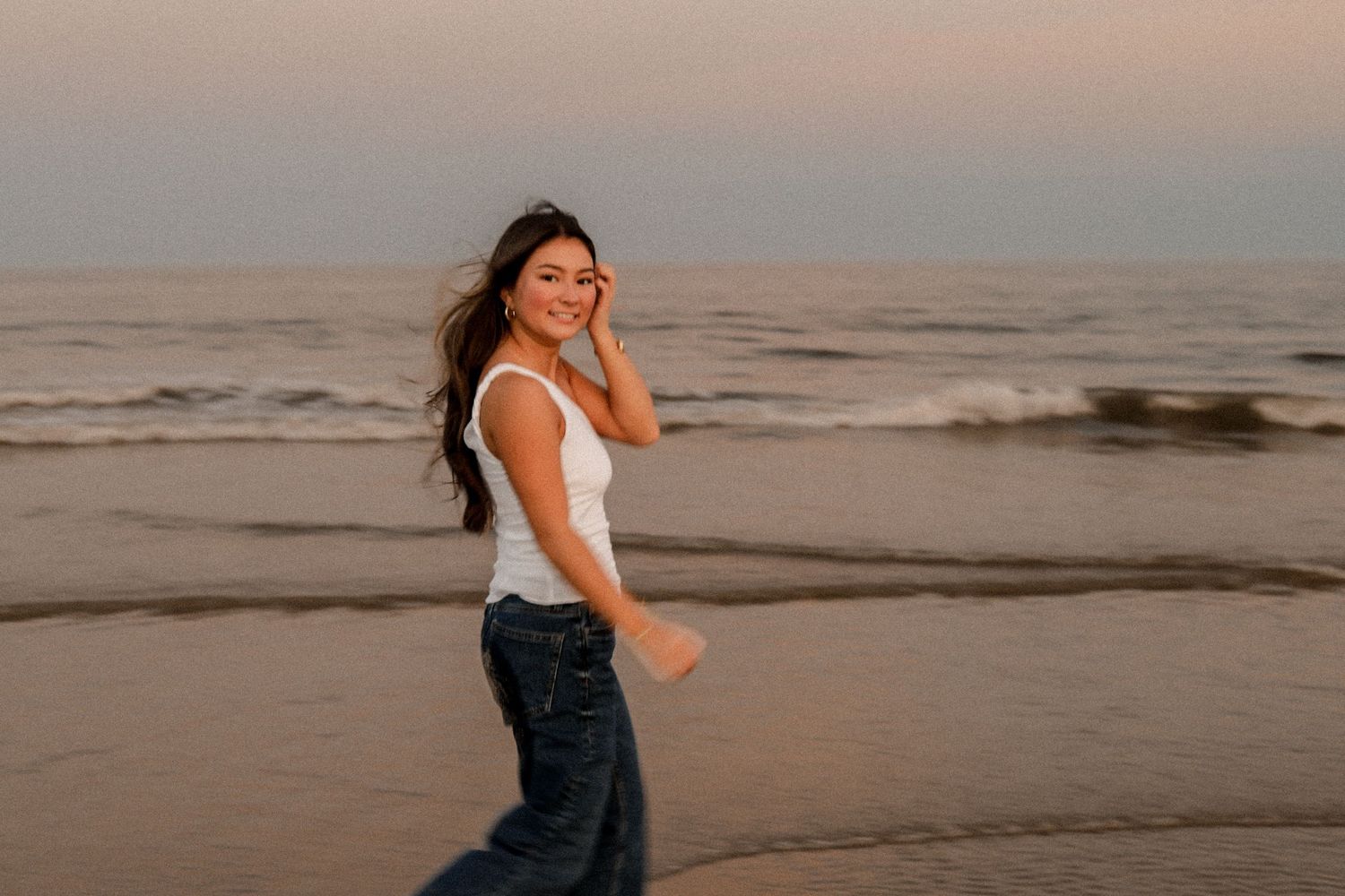 A person in white top and dark skirt dances joyfully on a sandy beach at sunset.