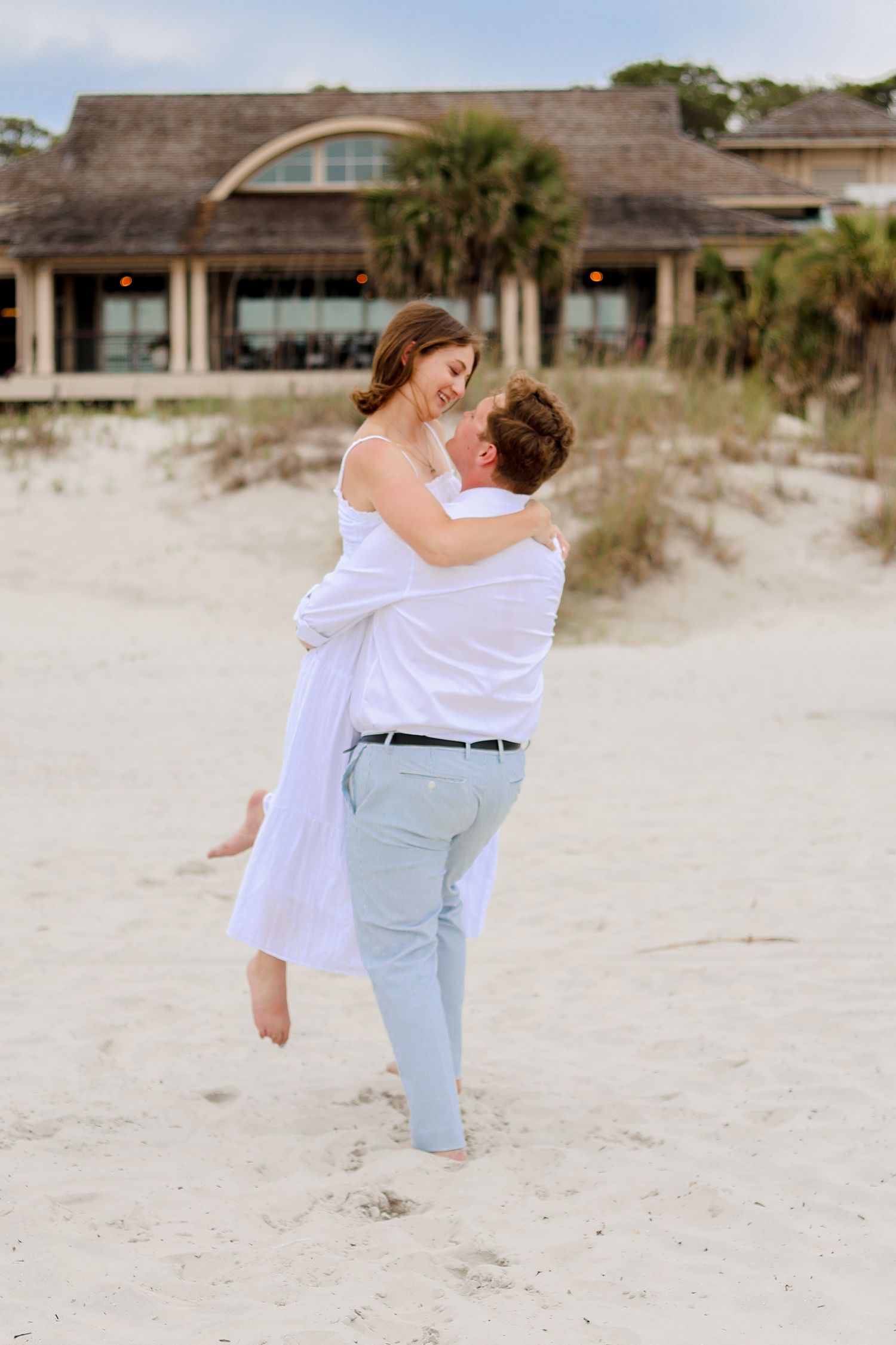 A romantic couple embraces while standing on a sandy beach with a thatched roof building in the background.