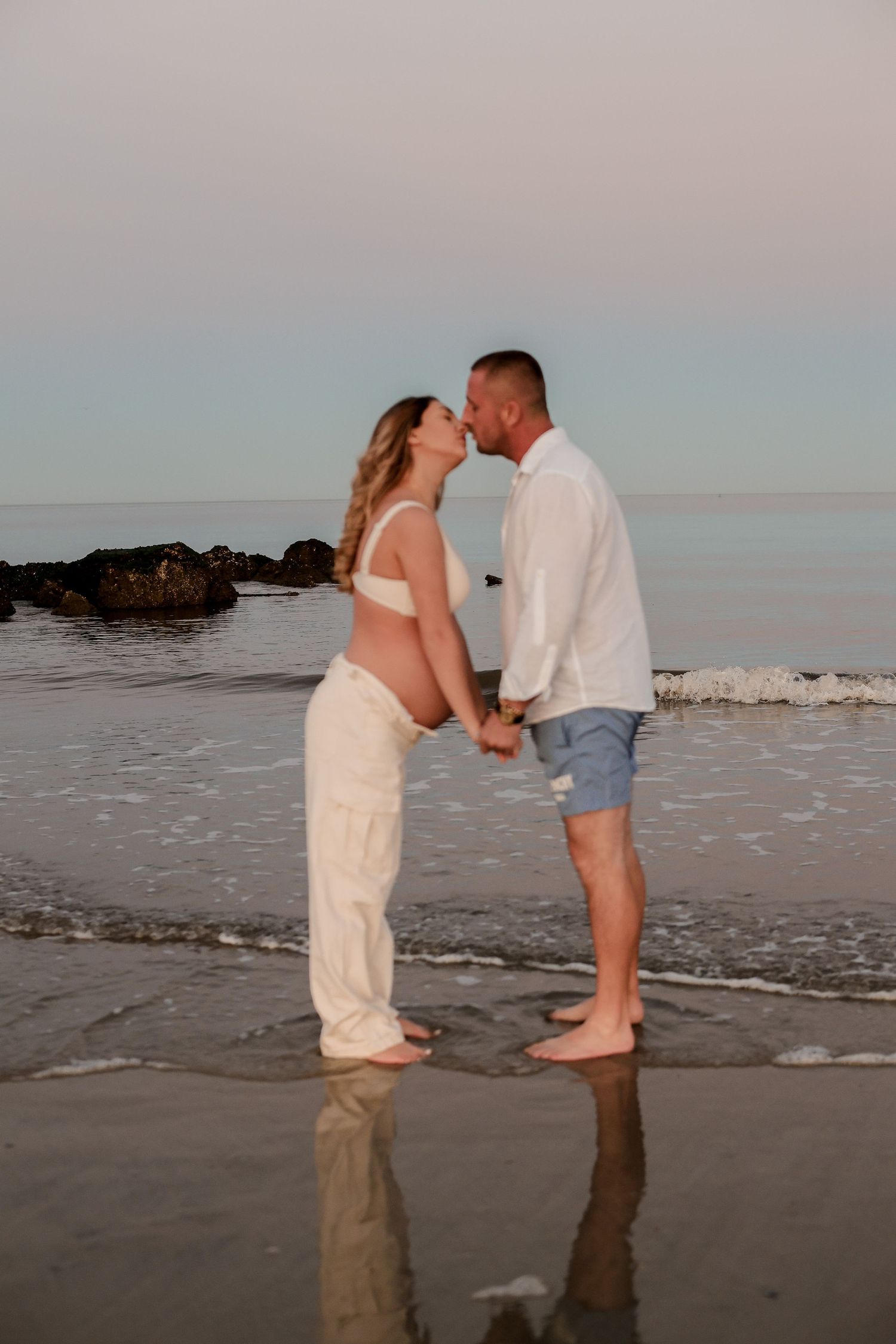 Couple stands in shallow ocean water during sunset, sharing an intimate moment on the beach.