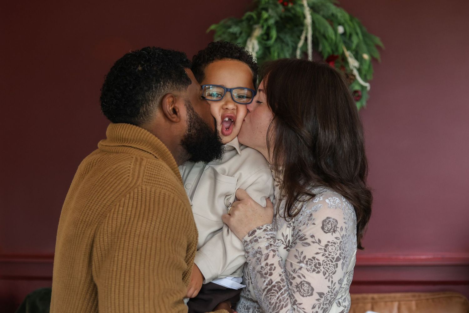A man and woman kiss the cheeks of a laughing young boy in glasses during a festive holiday moment indoors.