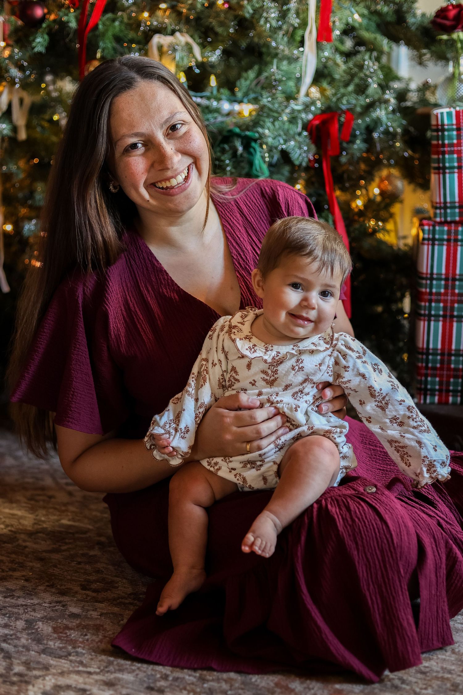 Smiling woman in a burgundy dress holds a baby in a floral outfit while sitting in front of a decorated Christmas tree and wrapped gifts.