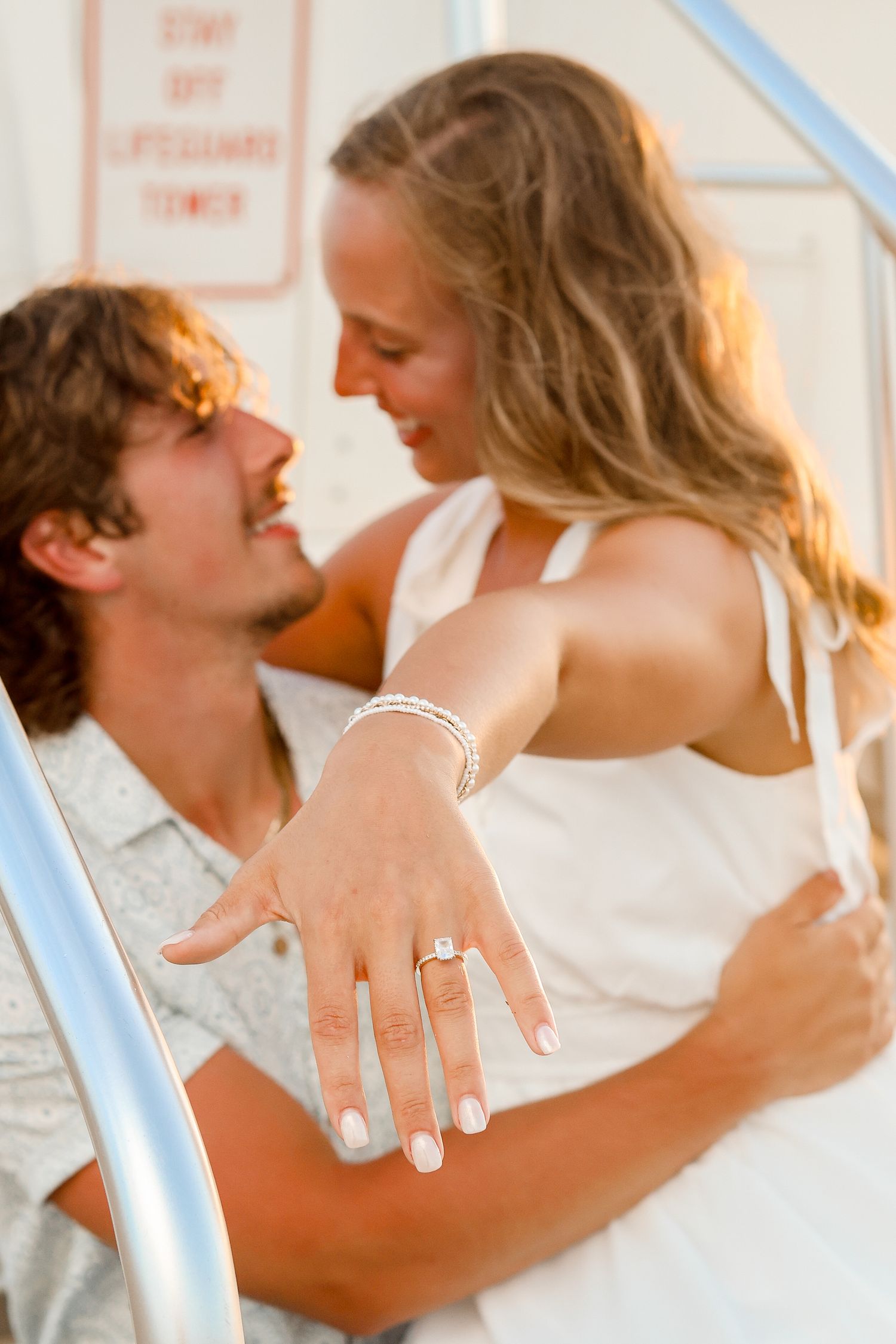 Couple showing off engagement ring while embracing in front of a lifeguard tower.