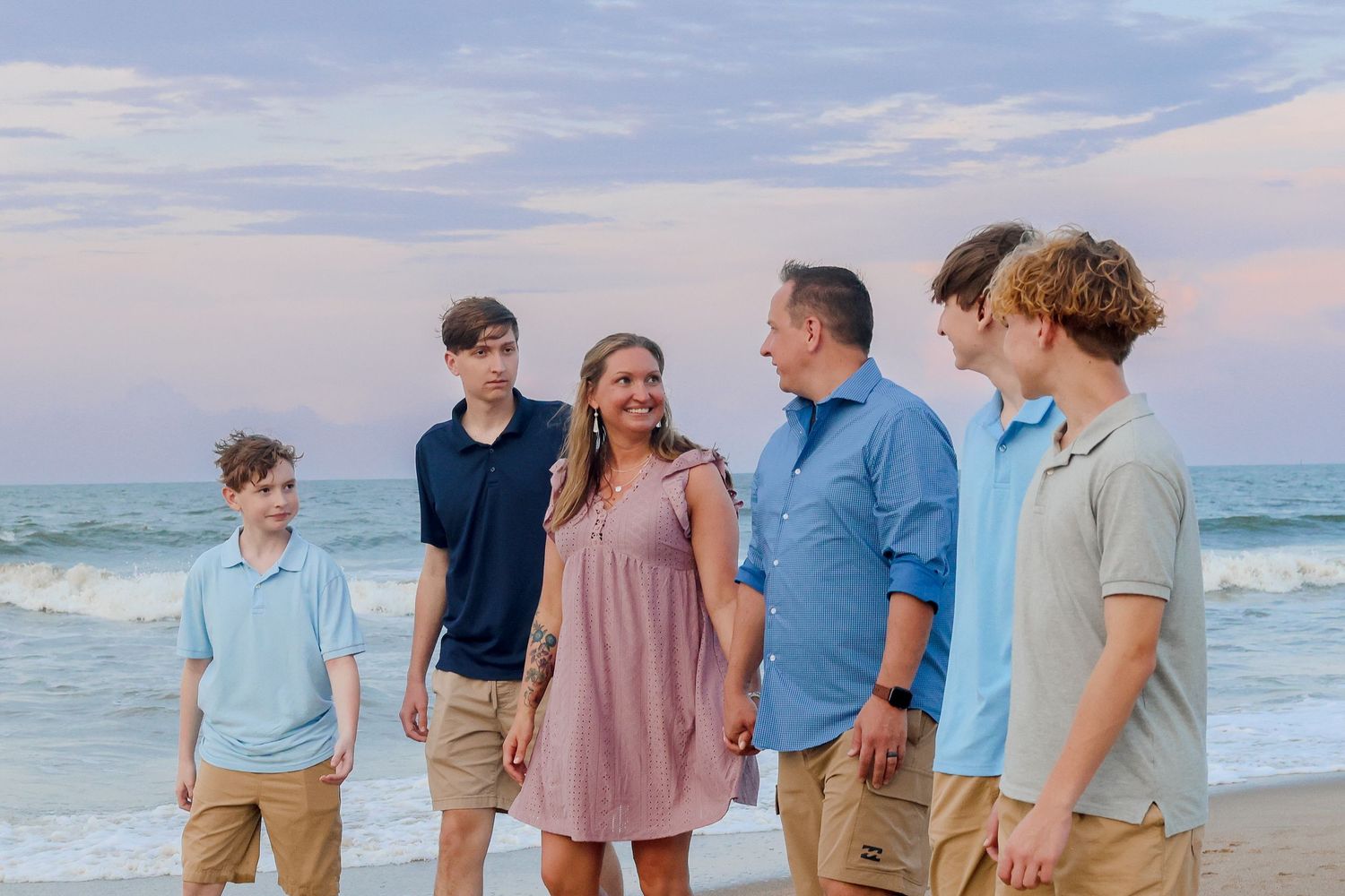 Family group wearing coordinated blue and neutral outfits poses together on beach at sunset.