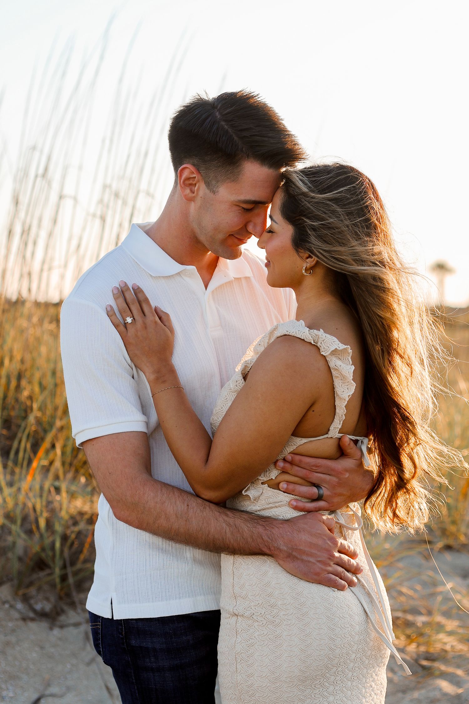 A romantic couple embraces on a beach at sunset with tall grass in the background.