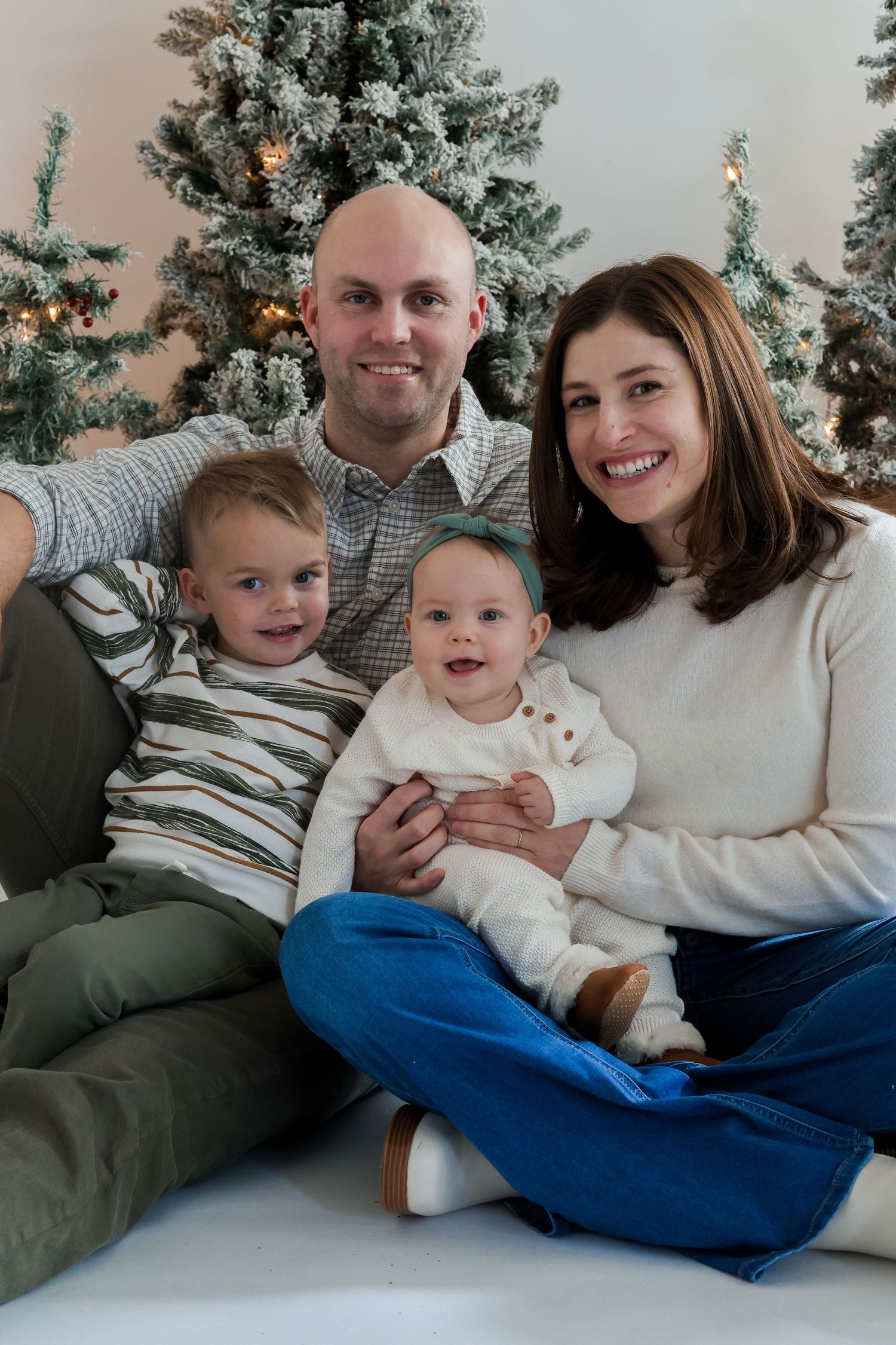 Casual family photo session with Christmas trees and twinkling lights in background.