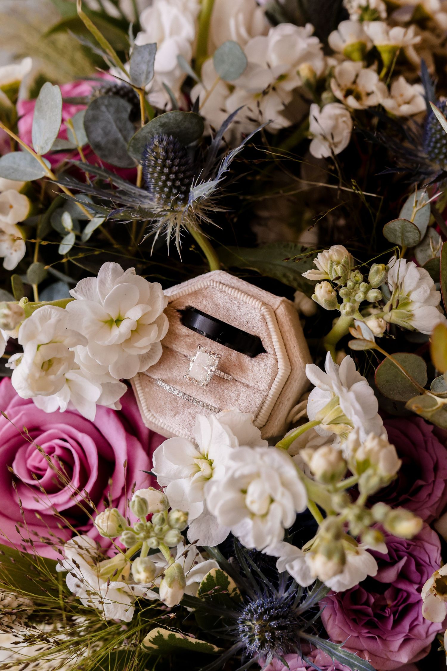 A wedding ring box nestled in a romantic bouquet with pink roses, white flowers, and blue thistle blooms.