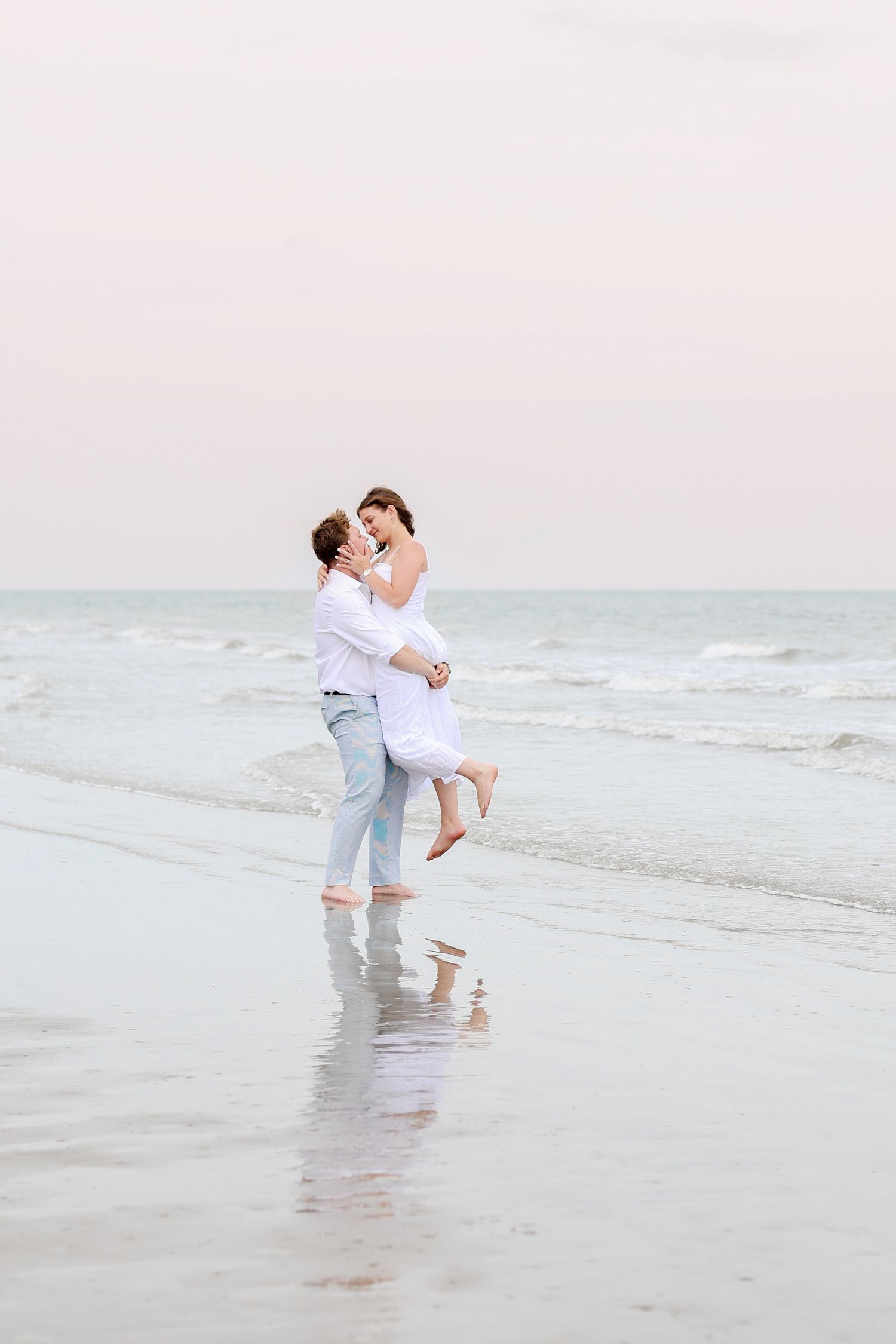 A romantic couple in white outfits embraces and dances together on a misty beach at sunset.