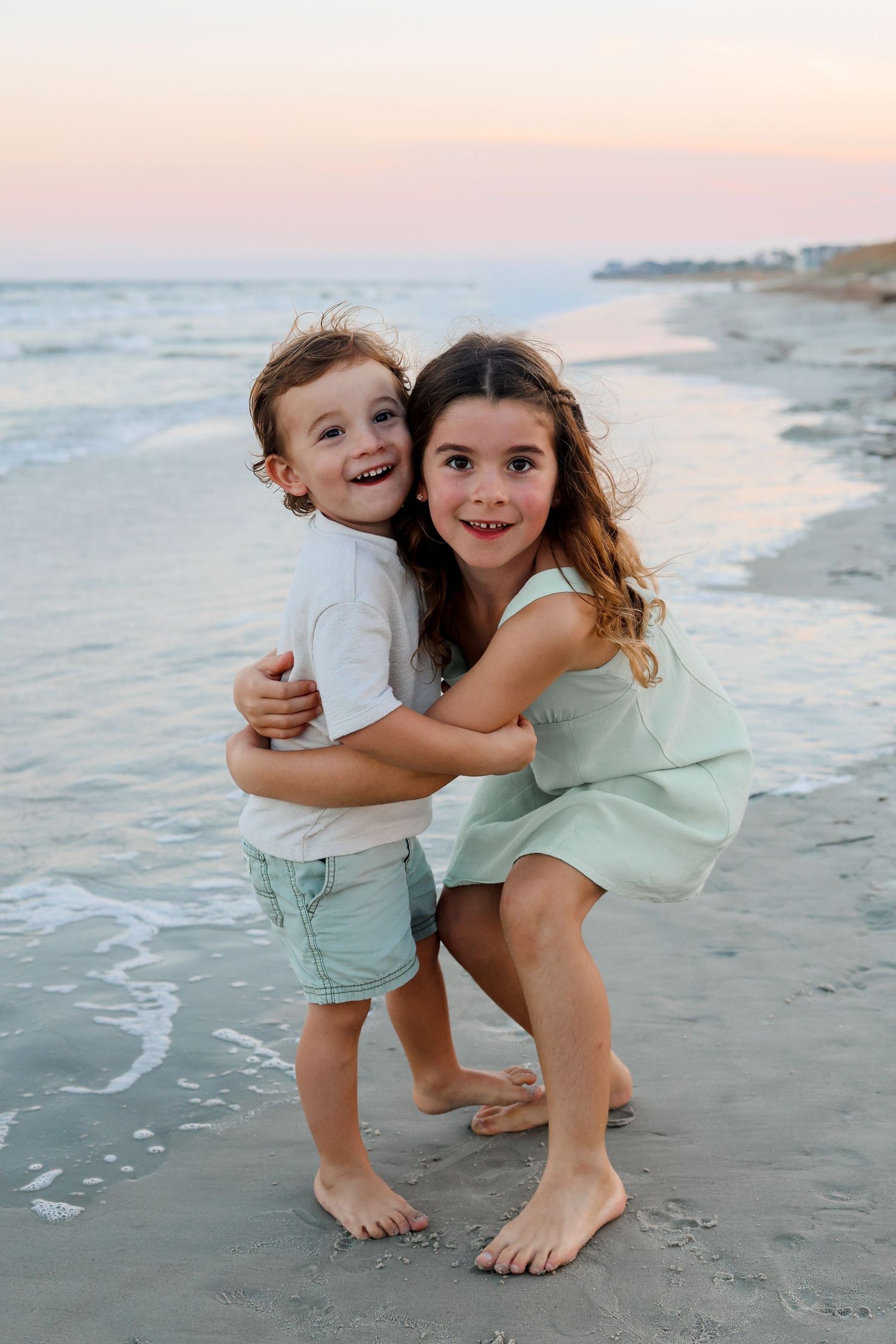Two children playing and hugging together on a sandy beach at sunset with ocean waves in the background.