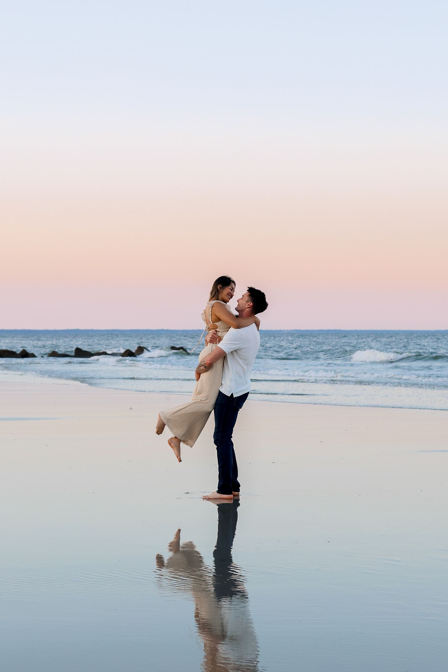 Couple sharing joyful lift pose on reflective beach water during sunset.